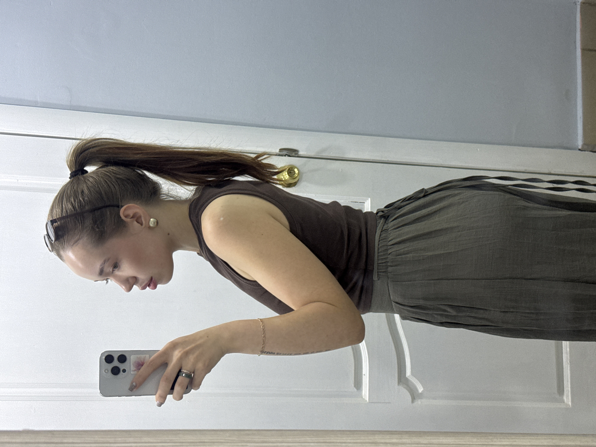 A woman with light skin is facing the lower-left corner, angled toward a mirror. She is holding a gray smartphone in her left hand, presumably taking a selfie. She has brown hair pulled back in a ponytail with a black hair tie, and she is wearing sunglasses atop her head. A small gold earring is visible in her left ear. She is wearing a brown sleeveless top and loose-fitting, dark green pants with a striped pattern along the side. A gold bracelet is on her left wrist. A ring is visible on the ring finger of her left hand. The background shows a white door with a gold handle, and a wall that is white at the bottom and light blue at the top. There is no indication that this image was taken in the city of Chaikovsky, Russia.