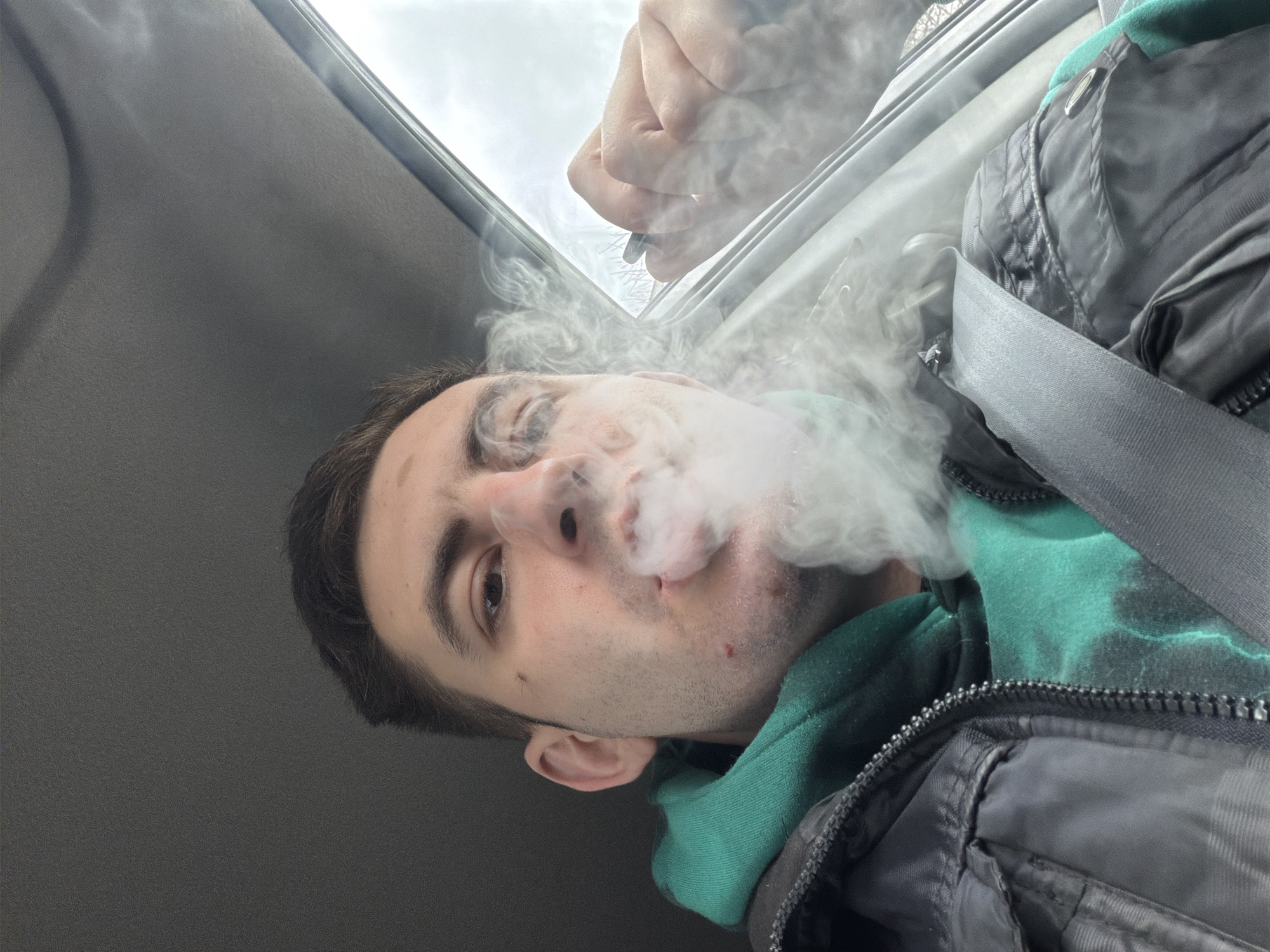 A man is in the driver's seat of a car, wearing a green hoodie and a black jacket with the seatbelt fastened. He is exhaling smoke, possibly from a cigarette or vape pen held by another person outside the open sunroof. The car's interior is gray. The location is not discernible from the image.
