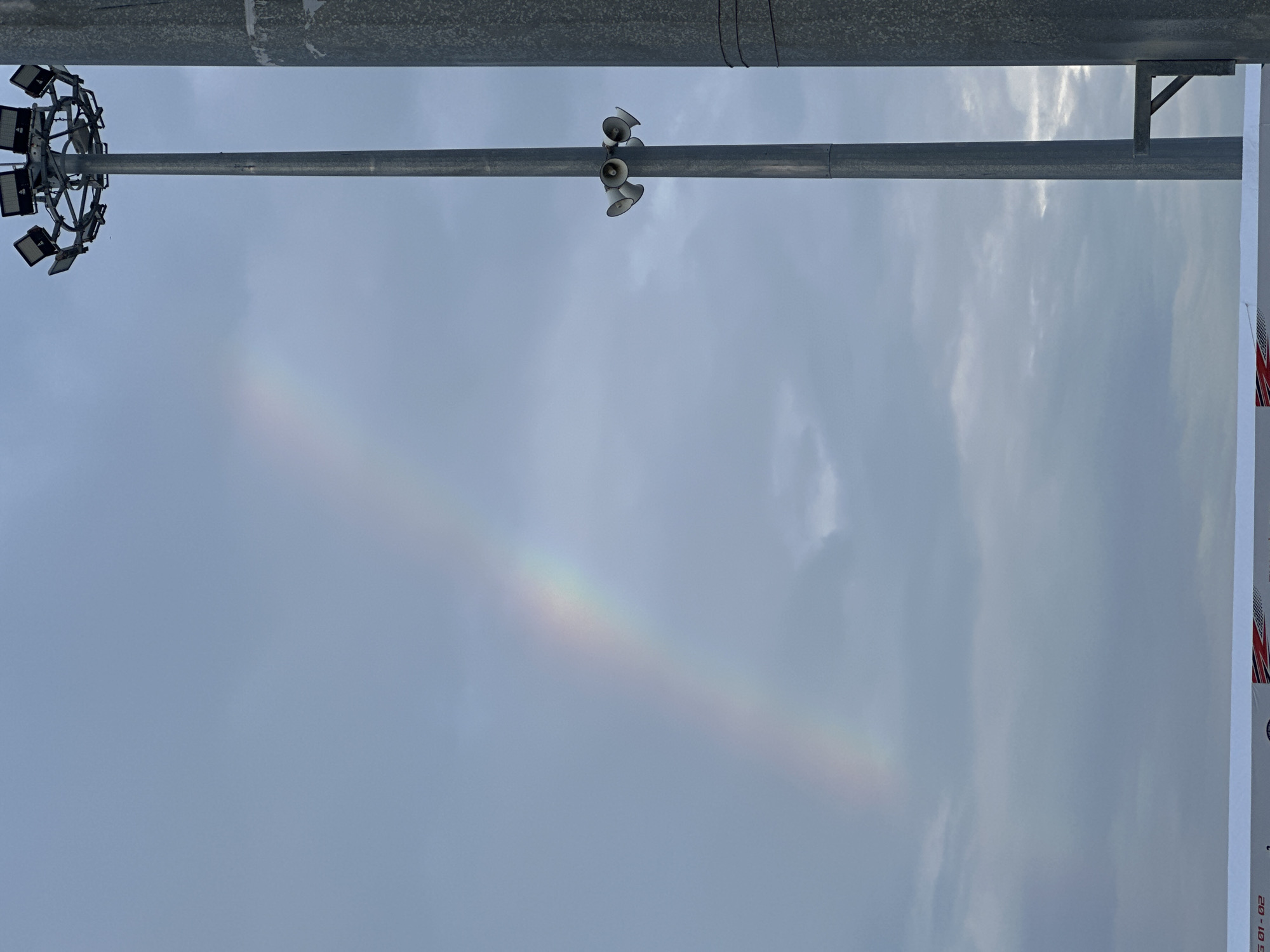 The image captures a serene outdoor scene, dominated by a light blue sky with scattered, soft white clouds. A prominent, thick grey metallic pole stretches horizontally across the upper portion of the frame.
Attached to the left side of this pole is a large, circular array of floodlights, featuring numerous black, rectangular light fixtures pointing downwards. Further along the pole, towards the center-right, a cluster of four grey, horn-shaped speakers is mounted, angled slightly downwards. On the far right, a metallic bracket supports the pole, connecting it to an unseen structure.
The most striking natural detail is a faint yet distinct rainbow, or possibly a sun dog, arcing diagonally across the lower half of the sky from the bottom-left towards the upper-right. Its colors, though subdued, are visible, particularly in the center of the arc.
On the extreme right edge of the image, a white banner or sign is partially visible. It features red and white stripes and a section of blue with white stars, strongly resembling a portion of the American flag. Below this, some dark text is visible, including "01-02" and possibly "20" above it, though other characters are obscured.
The overall impression is that of a daytime scene, likely after a period of rain, allowing for the formation of the rainbow. The presence of the floodlights and speakers suggests a public space, such as a stadium, park, or a large outdoor venue. The location context provided indicates this scene is in Nha Trang Ward, Vietnam.