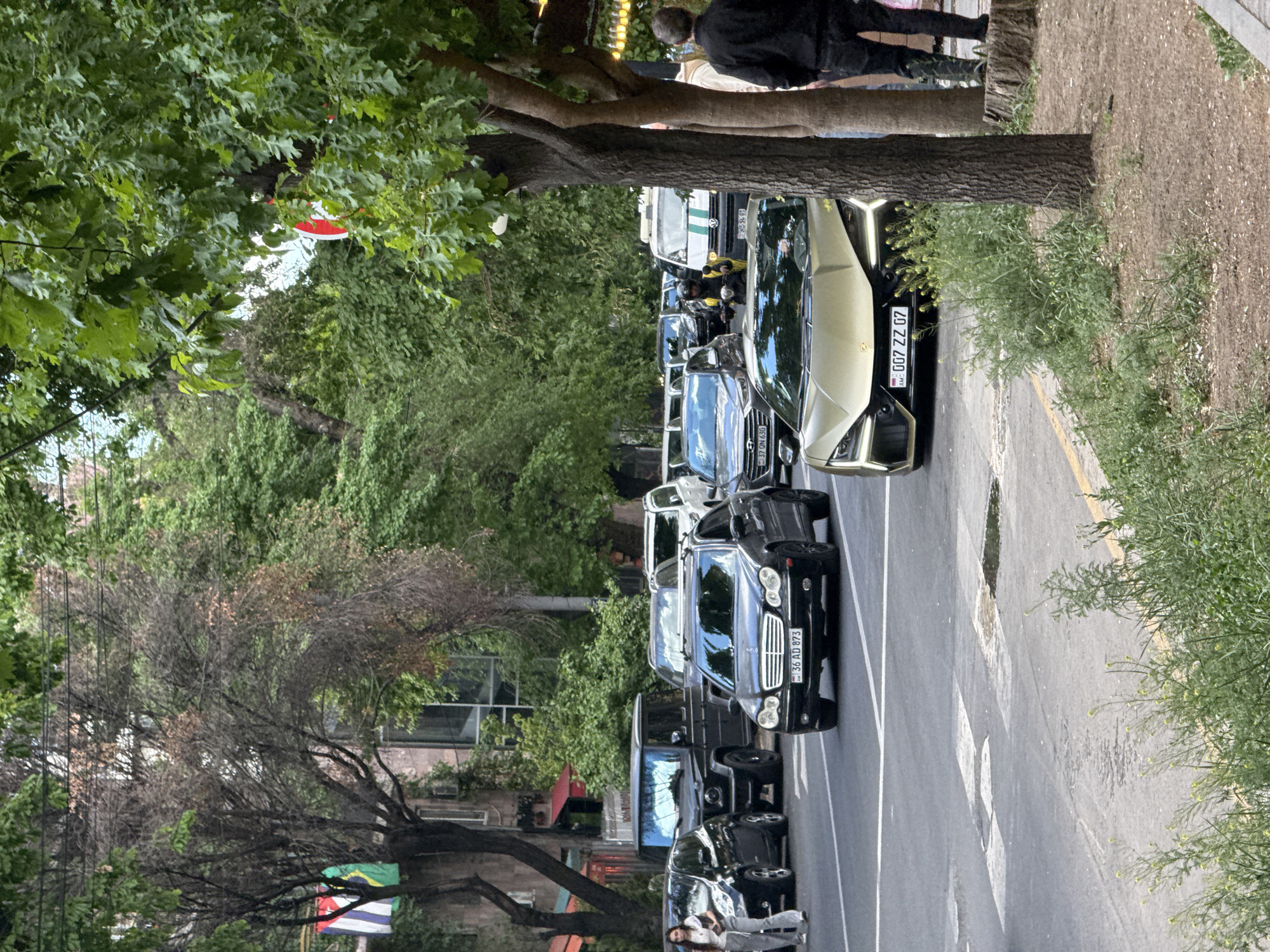 The scene is a street in Yerevan, Armenia, lined with trees and parked luxury vehicles. The camera captures a vertical view of the street, focusing on the cars and foliage along the roadside.
Several luxury cars are parked along the street, including a Lamborghini with a license plate that reads "007 ZZ 07" and a Mercedes-Benz with the license plate "36 AD 873." Other luxury vehicles are also parked along the street.
The street is lined with lush trees, whose foliage partially obscures the sky. Patches of greenery fill the roadside. A flag is visible in the background.
A few people are visible. A couple walks on the sidewalk. Another person stands near the trees.