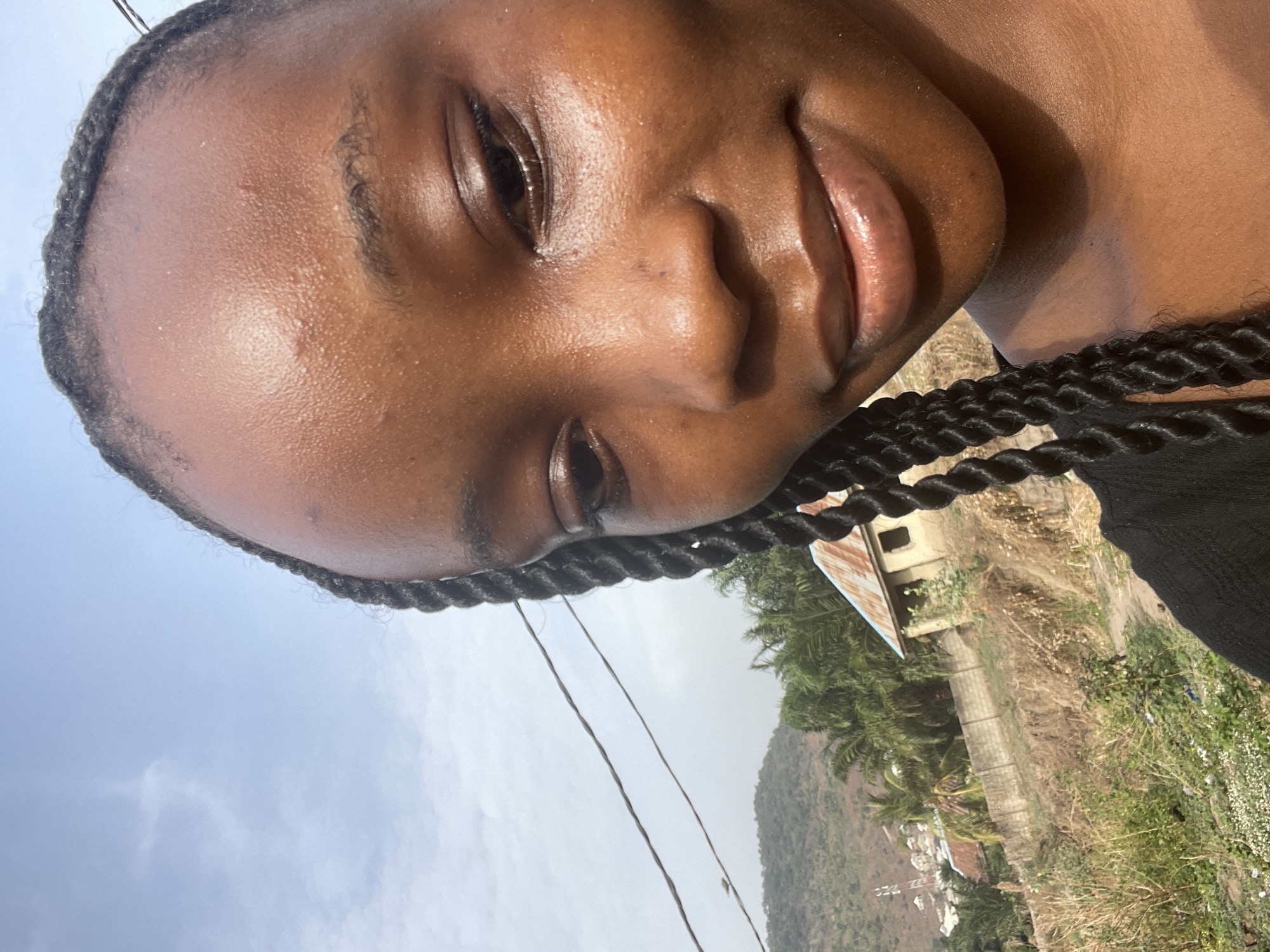 A close-up, rotated portrait captures a young Black woman, her face tilted to the right and filling the upper portion of the frame. She has a dark complexion, full lips adorned with a subtle gloss, and dark eyes looking directly at the viewer with a gentle, soft smile. Her hair is styled in neat, small braids that frame her face and extend over her shoulder, visible as a dark, textured mass on the right side of the image. A slight sheen is noticeable on her forehead, suggesting warmth or an oily complexion.
The background, visible to the lower right of her face due to the image's orientation, depicts an outdoor, rural or semi-rural scene. A bright, hazy sky with scattered clouds dominates the upper left. Below this, a verdant hilly landscape with varied green and brown vegetation, including palm trees, extends into the distance. In the mid-ground on the right, a small, simple building with a rusty corrugated iron roof stands amidst dry grass and shrubs. What appears to be a concrete path or low wall runs horizontally near the building. Overhead, thin power lines traverse the scene against the sky and distant hills. The lighting is bright, indicating a sunny day. No visible text is present in the image. The setting is consistent with a warm climate, possibly Lokoja, Nigeria.