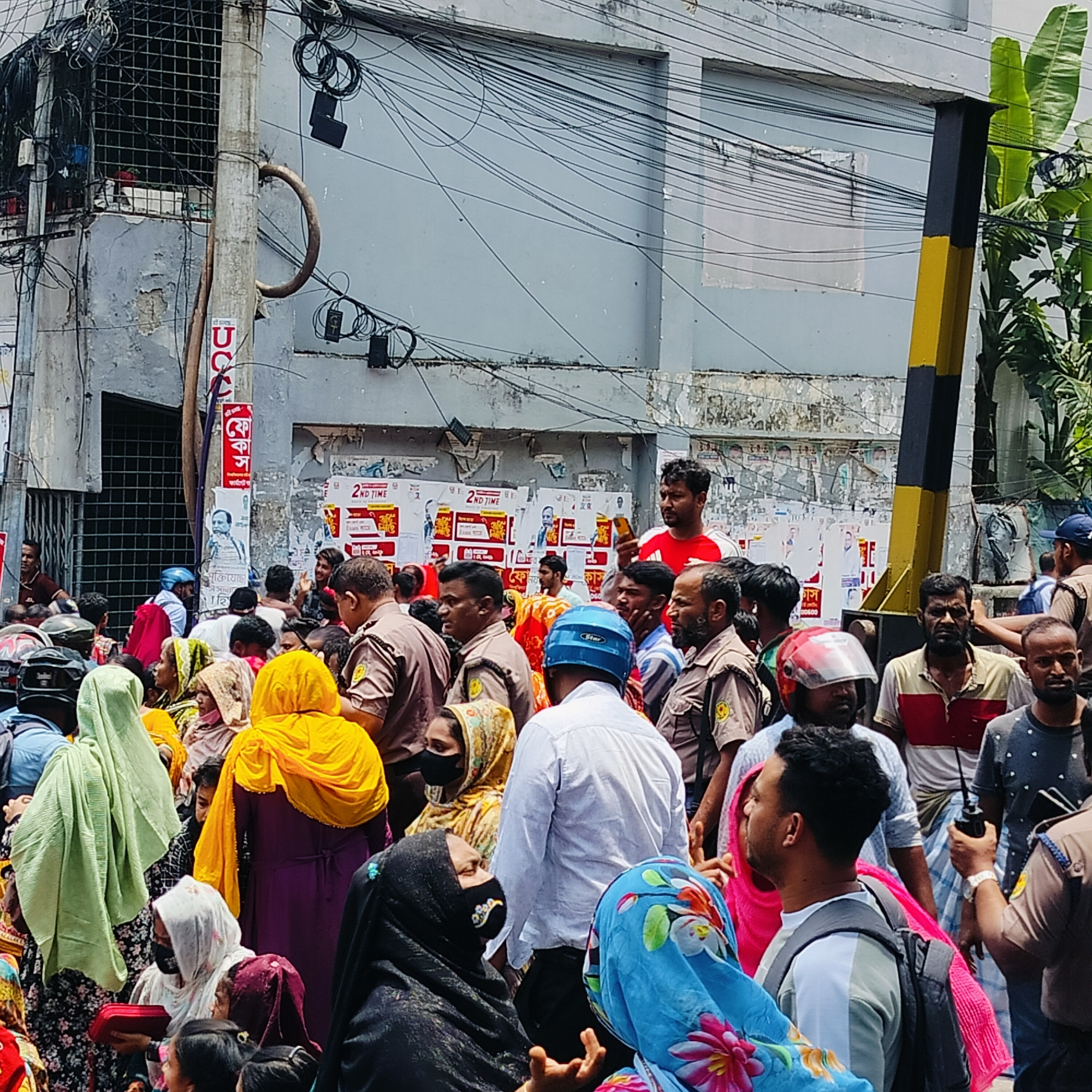The image depicts a densely packed crowd of people in what appears to be a street in Dhaka, Bangladesh. The majority of individuals are wearing garments of various colors and styles, with many women wearing hijabs or other head coverings. Several men are wearing uniforms, suggesting they may be police officers or security personnel. A man in a red shirt stands out slightly from the crowd.
The background features a building with multiple posters and wires running across its facade. A utility pole with black and yellow stripes is visible on the right side of the image.
The scene conveys a sense of activity and potential commotion in a densely populated urban environment.