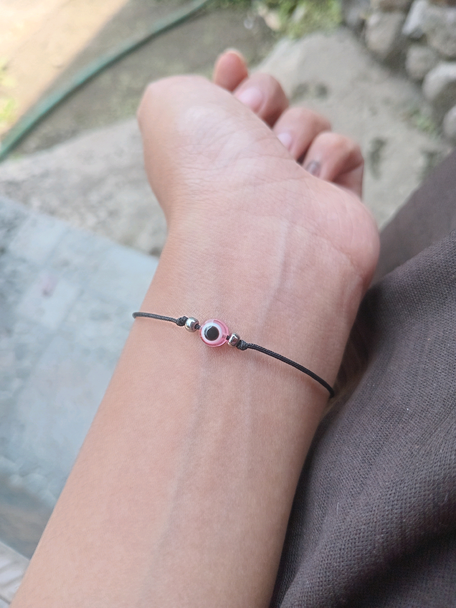 A close-up shot focuses on a person's left wrist wearing a thin black string bracelet. The bracelet features a pink evil eye charm, accented by two small silver beads on either side of the charm. The background is blurred, showing an outdoor environment with a concrete path and some greenery. The image was taken in Claket, Indonesia.