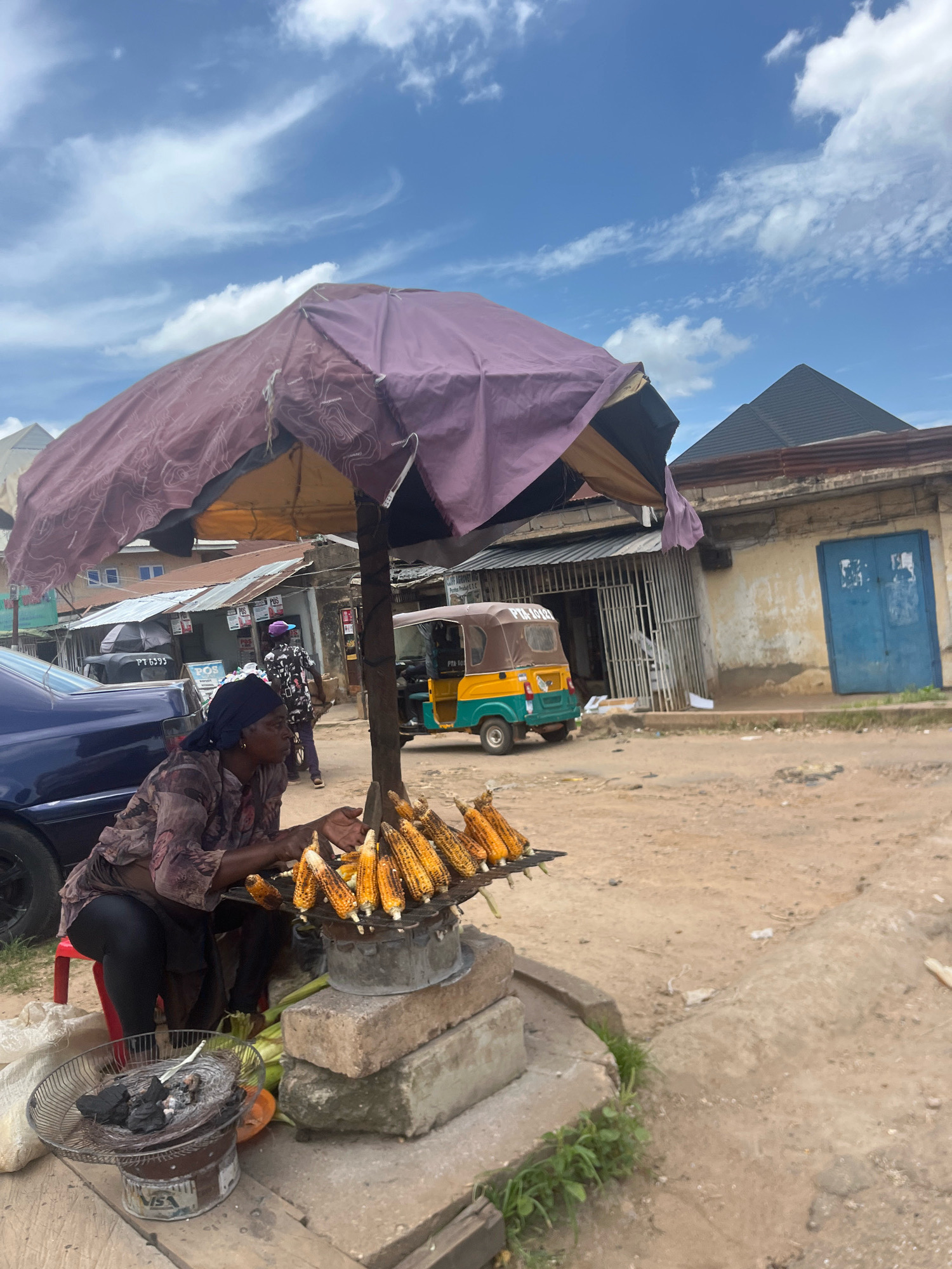 A street vendor is selling roasted corn under a makeshift awning in Jos, Nigeria. The vendor, a woman wearing a headscarf and a patterned top, is seated on a red stool next to a metal grill where several ears of corn are being roasted over charcoal. In front of the grill, a smaller charcoal brazier is visible. The scene appears to be on a dusty street, with buildings and a yellow and green auto-rickshaw in the background. The sky is blue with scattered clouds.
