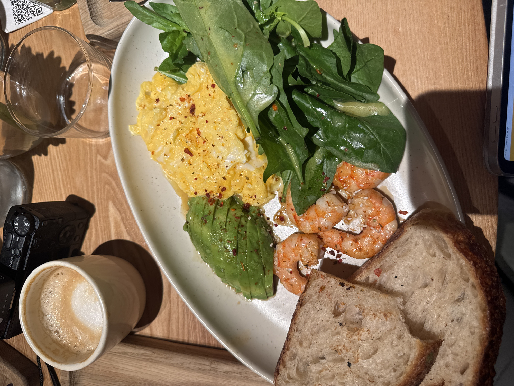 A high-angle shot reveals a light-colored, oblong ceramic plate containing a breakfast or brunch meal. The meal consists of scrambled eggs, sliced avocado, cooked shrimp, a green salad, and two slices of sourdough bread. Red pepper flakes are sprinkled on the eggs, avocado, and shrimp. The salad appears to be dressed with a light vinaigrette.
Adjacent to the plate, on the left side, is a cup of coffee with latte art, next to a black digital camera. A drinking glass is also visible in the background. The scene is set on a wooden tabletop. Based on visual clues and commonly available information, the location could be a cafe or restaurant in Yerevan, Armenia.