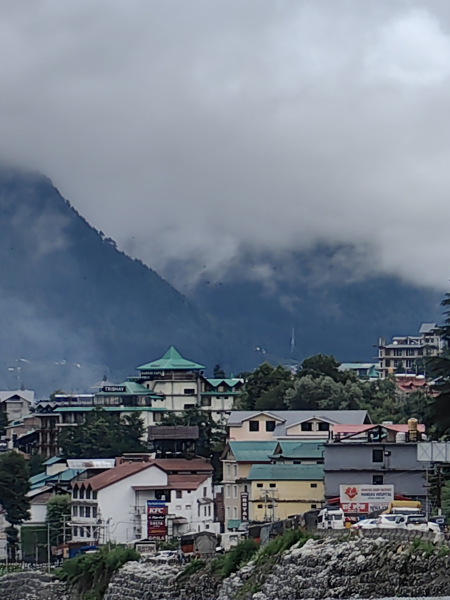 The image depicts a view of Manali, India, on a cloudy day. The foreground is dominated by a rocky embankment covered in green vegetation. Beyond this, a dense cluster of buildings forms the cityscape. Many of these structures have multi-story designs with pitched roofs, some appearing to be hotels or guesthouses. Notable signs visible on the buildings include "KFC," "COSTA," "TRIBHAV," "RADHIKA CAFE," and "MANGAV HOSPITAL." Several vehicles, including cars and what appear to be vans, are parked on the street level. The mid-ground shows a lush green hillside dotted with trees. In the background, a large mountain looms, its peak obscured by heavy clouds, creating a dramatic and somewhat somber atmosphere. The overall scene suggests a typical bustling town nestled in a mountainous region.