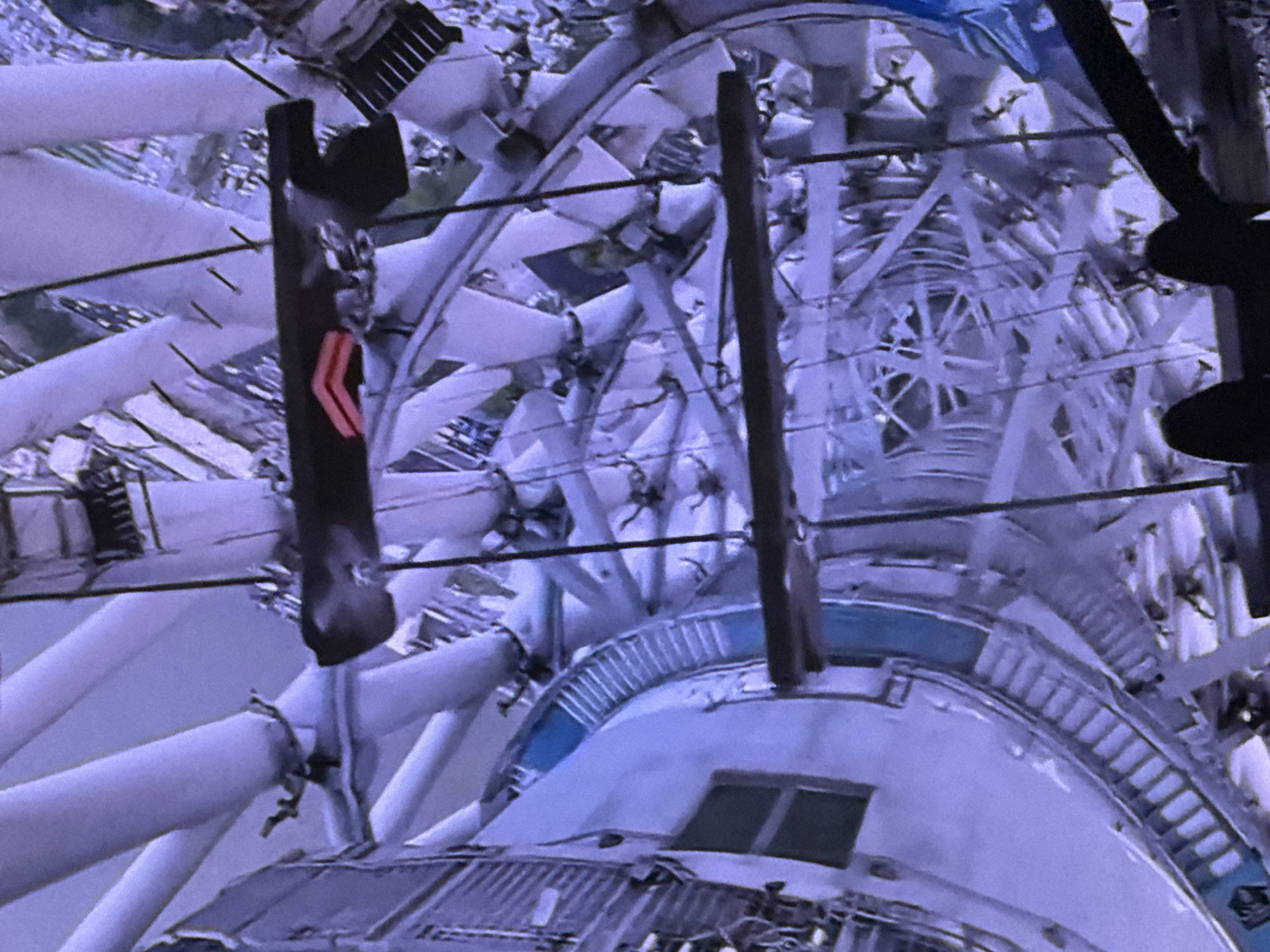 This is a tilted, medium-shot, full-color photograph taken indoors, looking down into the interior of the Ostankino Tower in Moscow, Russia. The tower's structural elements, comprised of large white cylindrical pipes and a complex network of metal beams and cables, dominate the scene. A section of what appears to be a circular staircase with blue treads is visible on the left side of the frame. Two dark, horizontal beams stretch across the middle ground, one appearing to have a red object attached. The background is a blur of similar structural elements, suggesting significant height and depth within the tower. The lighting is uneven, creating shadows and highlights across the metallic and pipe-like surfaces. There are no visible people or animals in the frame.