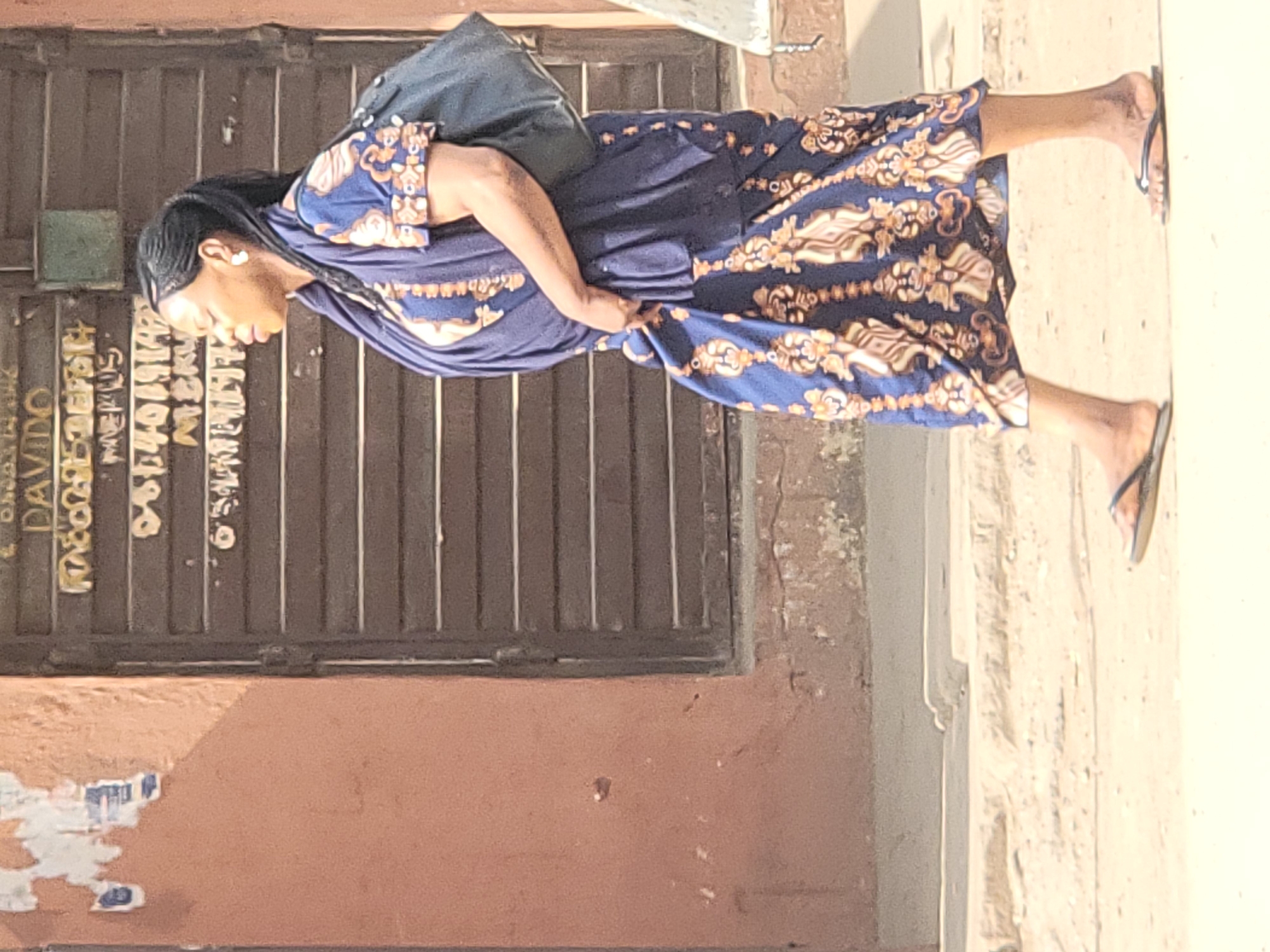 An outdoor, eye-level shot captures a woman walking or standing on a bright, sandy-colored ground next to a building in Maiburiji, Nigeria. She is wearing a long, blue dress with a distinctive brown, gold, and cream patterned hem and sleeve cuffs. Her hair is styled in dark braids, and she wears white heart-shaped earrings. On her back, she carries a dark-colored bag. Her feet are visible, clad in simple black and brown flip-flops. Her gaze is directed downwards, and her right hand appears to be adjusting or holding the fabric of her dress.
The background consists of a dark brown or black metal gate or door with vertical slats. Below the gate is a reddish-brown wall, showing some signs of wear and peeling plaster in the lower left. Graffiti is visible on the dark gate; notably, the word "DAVIDO" is clearly legible in white lettering, oriented vertically near the left edge. Other white or light-colored markings and symbols are present on the gate but are mostly indistinct. The bright sunlight suggests it is daytime.