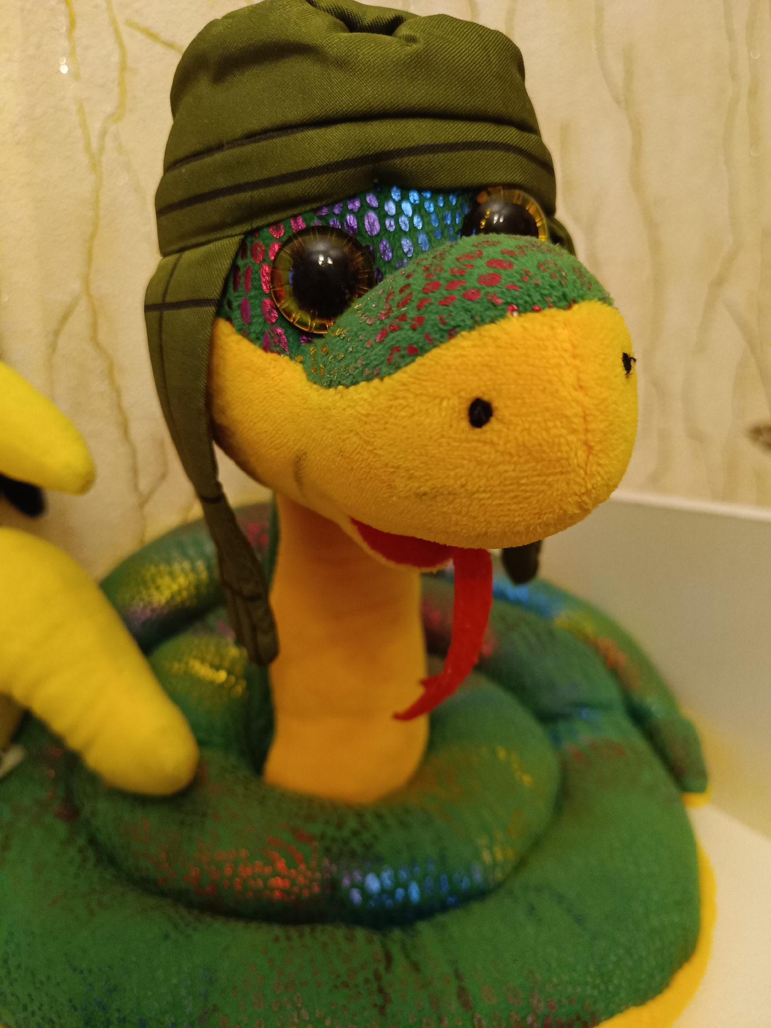 A plush snake toy wearing a green hat is coiled on a surface. The snake is primarily green with yellow and red accents, featuring large, glossy black eyes. The hat is a darker shade of green with horizontal stripes. The background appears to be a patterned wall, possibly in a home setting. There is no information to confirm that the image was taken in Tobolsk, Russia.