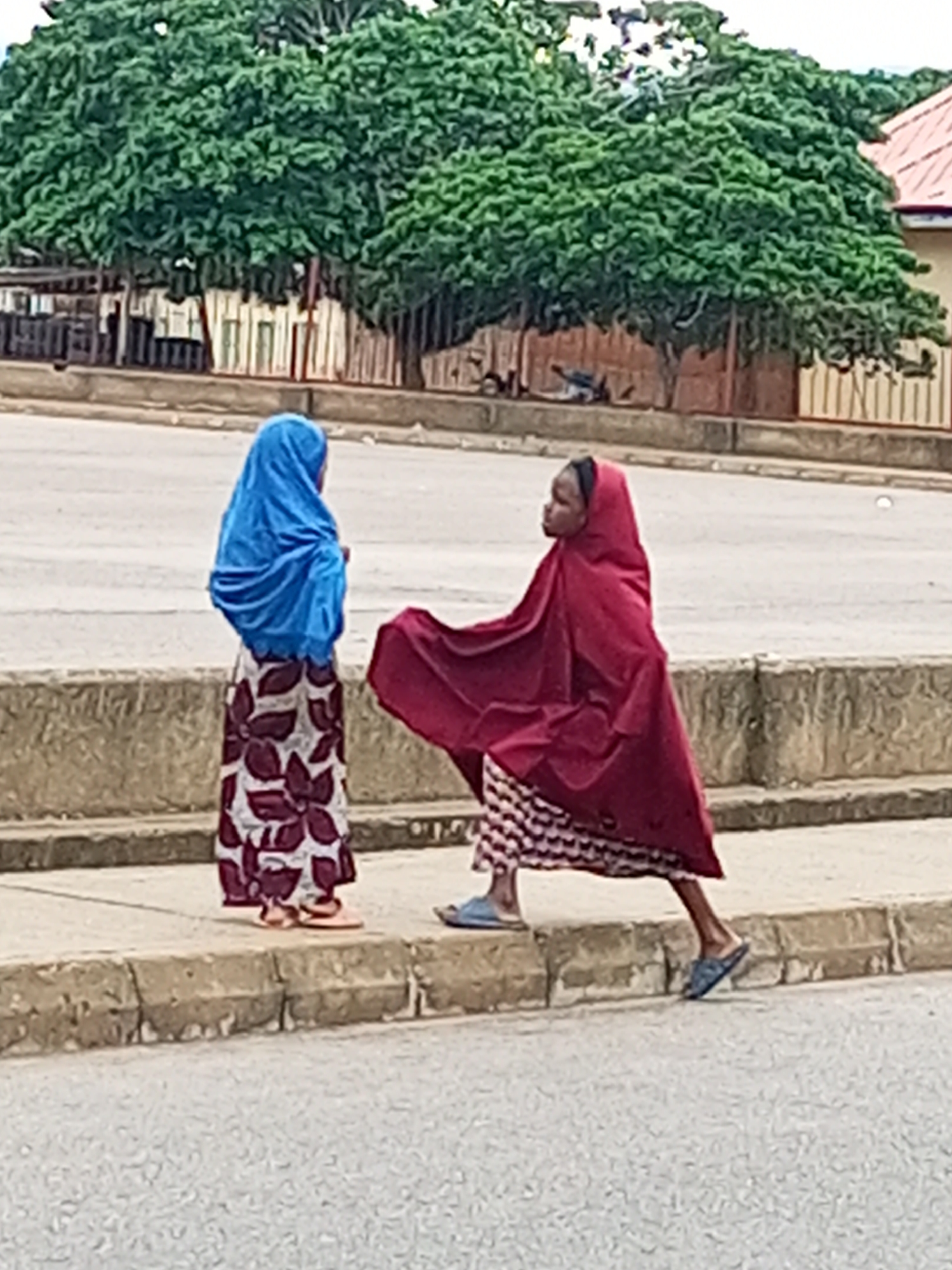 Two young girls are standing on a concrete curb in front of a paved surface, likely a road. The girl on the left is wearing a bright blue hijab and a long skirt with a large floral pattern in white and dark red. She is facing to her right and looking towards the other girl. The girl on the right is wearing a maroon hijab and a patterned skirt in white and dark red, with a checkered design at the hem. Her maroon hijab is draped around her, covering her shoulders and extending to her waist. She is facing to her left, appearing to be interacting with the other girl. Both girls are wearing sandals. Behind them, lush green trees with dense foliage create a backdrop, and in the far background, a building with a red-tiled roof is visible. The setting suggests an outdoor urban environment in Pantame, Nigeria.
