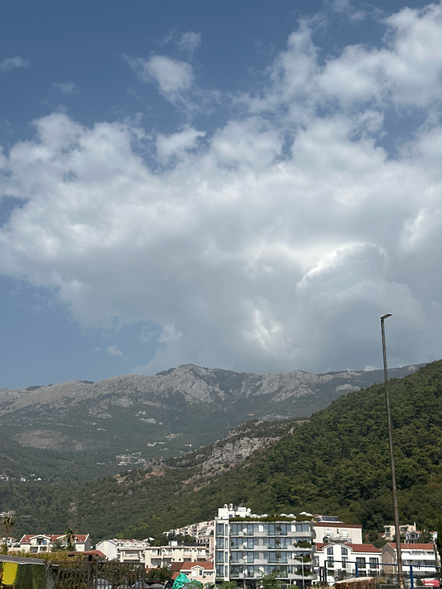 The image captures a panoramic view of Budva, Montenegro, under a partly cloudy sky. Dominating the upper portion of the frame are large, voluminous clouds set against a clear blue sky. Below the clouds, a series of rugged, greyish-brown mountains stretch across the horizon. The lower slopes of these mountains are covered in dense, dark green vegetation, indicative of a forested area.
In the foreground, a modern, multi-story building with a white facade, large windows, and a flat roof adorned with what appear to be white umbrellas or coverings, stands prominently. The building has a sleek, contemporary design. Adjacent to it, several other buildings, mostly residential or small hotels, are visible, characterized by white and terracotta-colored roofs. A tall, dark pole with a modern lamp fixture stands to the right of the frame, slightly obscuring the mountain view. The scene suggests a coastal town nestled between mountains and the sea, with a mix of natural landscape and urban development. There are no visible people in the image.