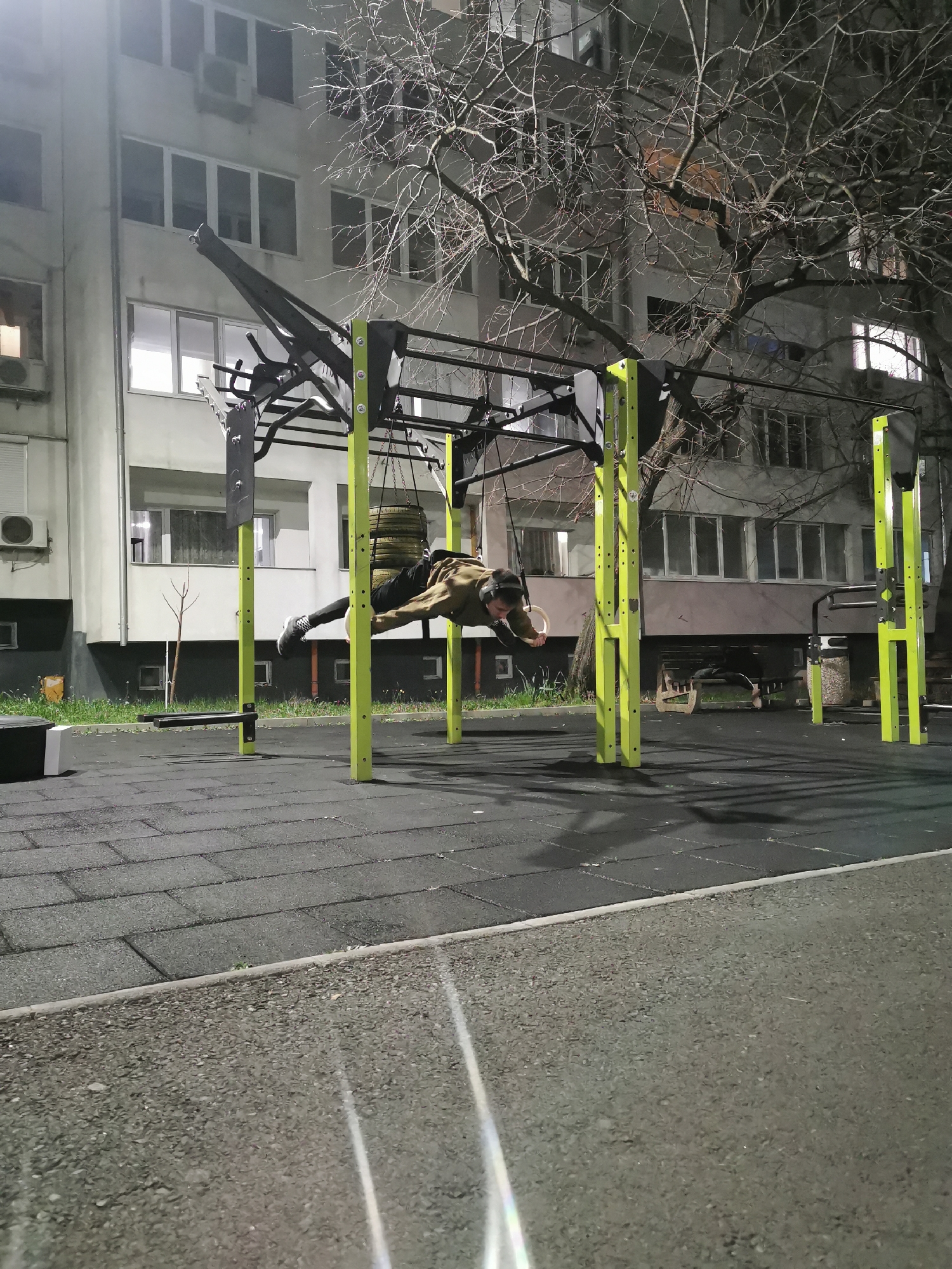 An individual is suspended mid-air on gymnastic rings attached to a yellow and black outdoor fitness structure. The person wears dark pants, light-colored outerwear, and dark shoes. Behind the fitness structure, a multi-story building with windows is visible. A large tree with bare branches is also in the background. The ground is paved with interlocking rubber tiles, transitioning to a paved surface in the foreground. The scene appears to be illuminated by artificial light, suggesting it is nighttime. Based on the architecture and style of the surrounding environment, the location is likely in a residential area of Burgas, Bulgaria.