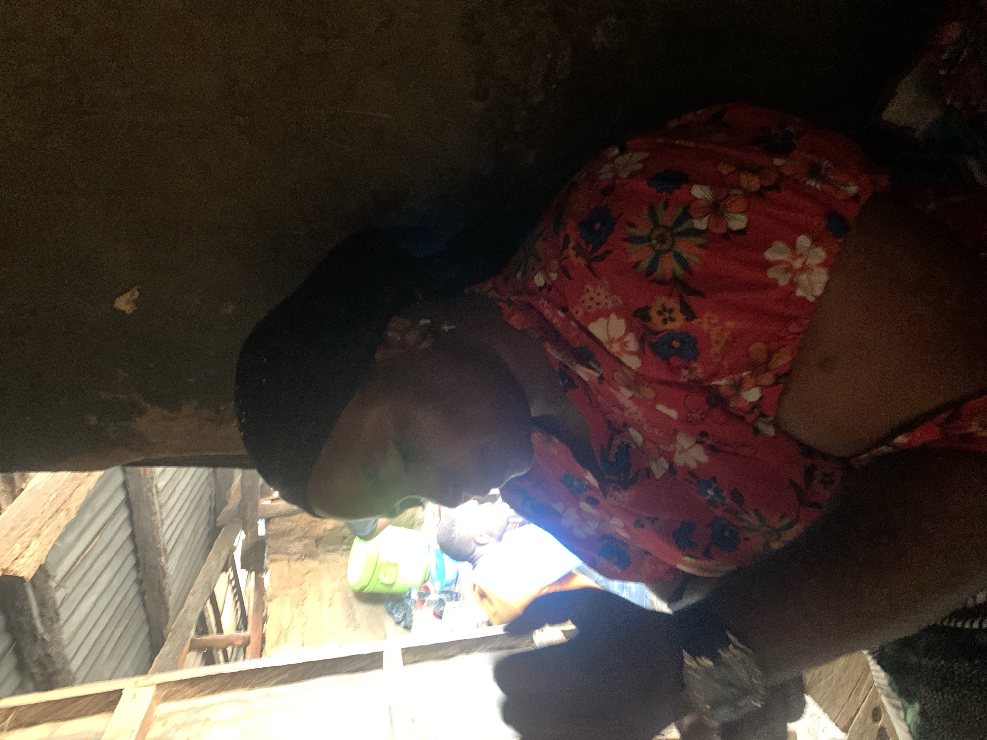 An African woman wearing a black headscarf and a red floral-patterned shirt is shown in profile, looking down and to the left. She has dark skin and appears to be in an indoor setting with natural light streaming in from the left. Behind her, another person in a white shirt is visible, along with some colorful plastic containers. The background appears to be a rustic structure with a corrugated metal roof and wooden beams.