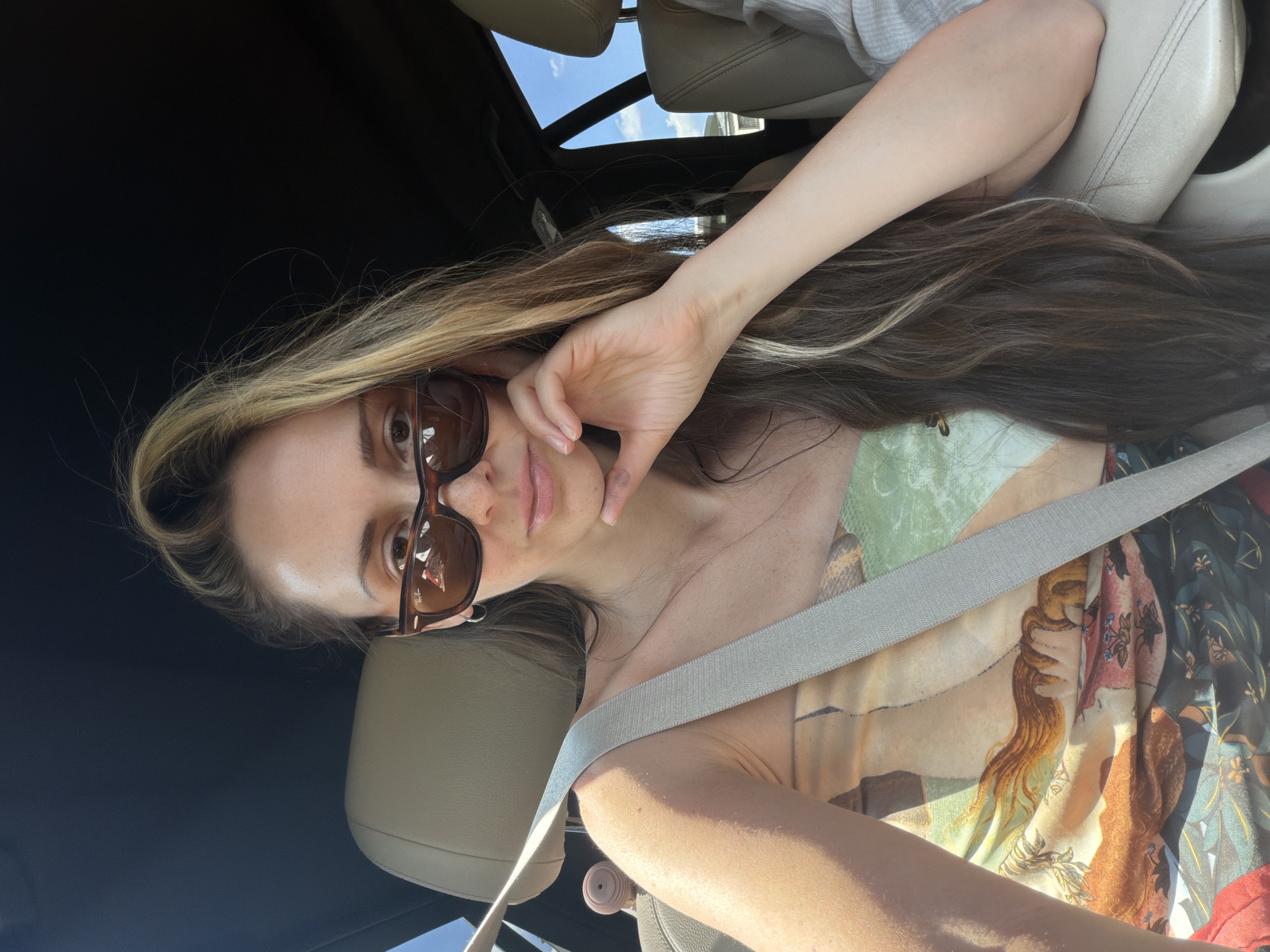 A woman with long, brown and blonde hair, wearing sunglasses and a patterned strapless top, is sitting in the driver's seat of a car. She is looking at the camera and resting her chin on her hand. A seatbelt is across her chest. The interior of the car is visible, including the beige leather seats and the black headliner. Outside the car window, the sky is blue with some clouds. The scene appears to be outdoors on a sunny day. There is no indication of the location being Moscow, Russia.