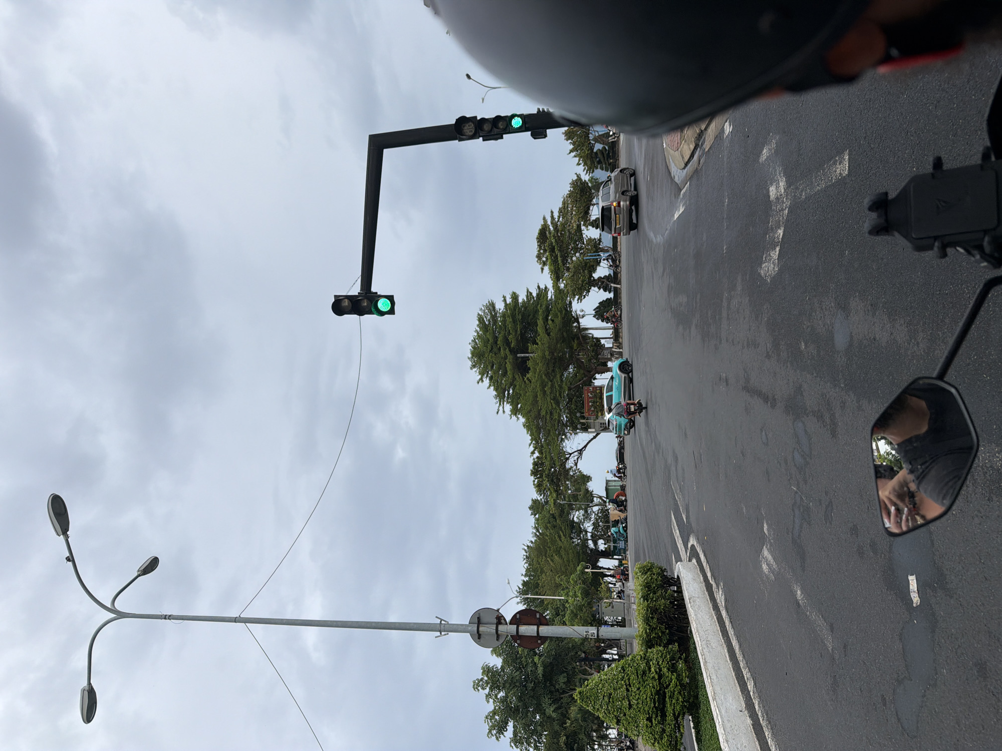 The image depicts a street scene in Nha Trang, Vietnam, on a cloudy day. The perspective is from someone riding a motorcycle, as evidenced by the rearview mirror showing a reflection of the rider's face and helmet. In the foreground, the asphalt road is visible with white lane markings.
A traffic light is prominent, with the green light illuminated. It is mounted on a dark metal structure extending over the road. Further down the road, a silver minivan is in motion, and a blue car is parked on the right side. Several motorcycles and scooters are also present on the road, with some riders visible.
To the left of the road, there is a well-maintained green bush and a white concrete curb. A tall street lamp with multiple lights stands behind this area. In the background, lush green trees line the horizon, suggesting proximity to a park or coastal area. The sky is overcast with gray clouds.