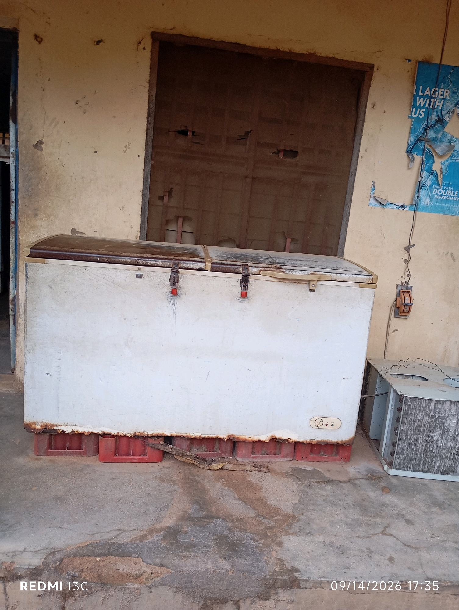 The image shows a weathered chest freezer situated outdoors on a concrete surface. The freezer is white with a dark-colored lid and appears to be supported by red plastic crates. To the right of the freezer, a disassembled air conditioning unit is visible, also on the concrete. In the background, a large, rustic-looking door is set into a building with beige, textured walls. A torn poster advertising "LAGER WITH QU" is affixed to the wall on the right. The ground is dusty and shows signs of wear and tear. The overall scene suggests an outdoor storage or abandoned area. The timestamp indicates the image was taken on September 14, 2026, at 17:35. The bottom left corner displays "REDMI 13C", identifying the phone model used for the photograph. There are no people or animals in the frame.