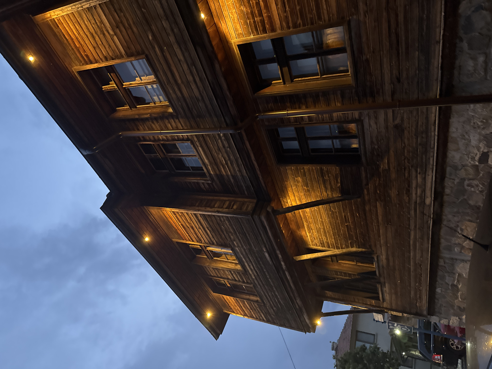 An exterior, low-angle, medium shot captures a two-story wooden building at dusk in Sozopol, Bulgaria. The building's facade is constructed of dark, horizontal wooden planks, some of which appear weathered. The lower portion of the building has a foundation made of irregular, rough-hewn stones. Several windows are visible on both levels; they are dark and reflect the warm glow of external lights. Small, warm-toned spotlights are affixed to the eaves of the building, illuminating the wooden siding.
To the left of the main building, a sliver of another structure with a tiled roof and lighter colored walls is visible, along with trees. In the foreground, the wet ground suggests recent rain, and the glint of light on the surface is evident. A dark-colored vehicle is parked on the street to the left, with a red vehicle partially visible behind it. A blue sign with white symbols and a white pole are also present near the parked vehicles. The sky above is a dusky blue with scattered clouds. The overall atmosphere is one of quiet evening in an old European town.