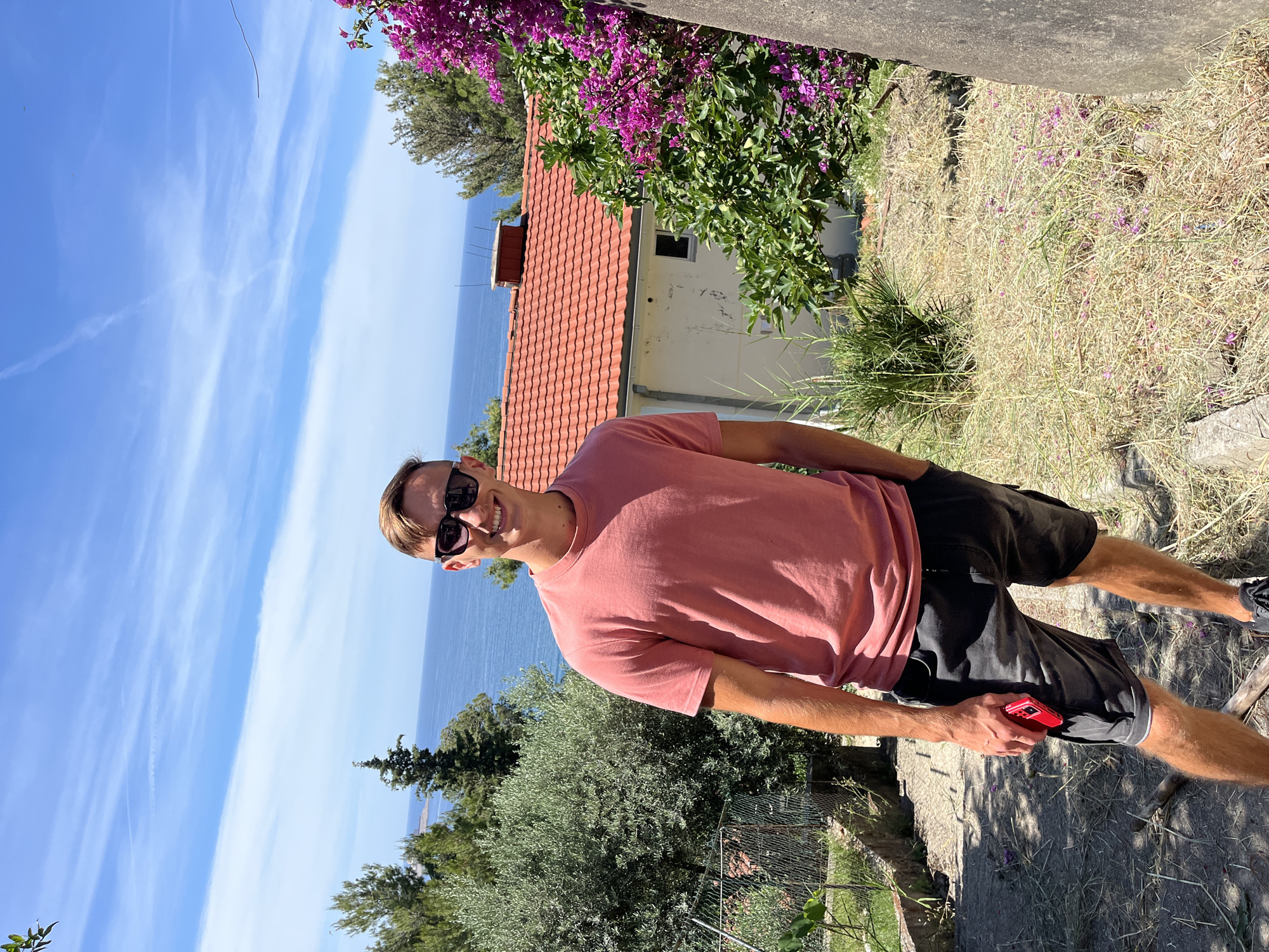 In Šušanj, Montenegro, a young man stands on a dirt path facing the camera, wearing sunglasses, a coral t-shirt, and dark shorts. He holds a red smartphone in his right hand. Behind him, a house with a red-tiled roof is visible, surrounded by green foliage and a vibrant pink flowering bush. The Adriatic Sea stretches out in the background under a bright blue sky dotted with white clouds. The ground is covered with dry grass and stones, and a metal fence runs along the left side of the path.