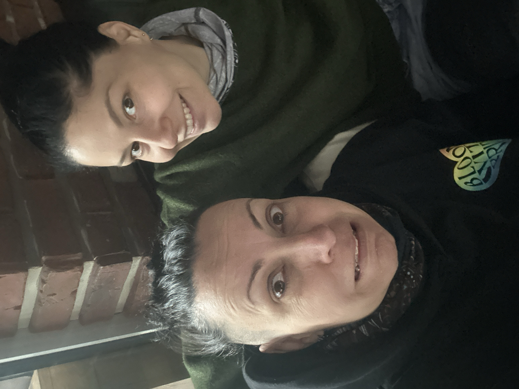 Two light-skinned women are posing for a selfie. One woman is wearing a green shirt with a gray scarf underneath, and the other is wearing a black sweatshirt with a heart-shaped rainbow-colored emblem. The emblem features the words "Blood. Sweat. Respect" in a circular pattern. The background shows brickwork, suggesting an indoor setting with a brick wall, possibly a cafe or apartment in Sofia, Bulgaria.