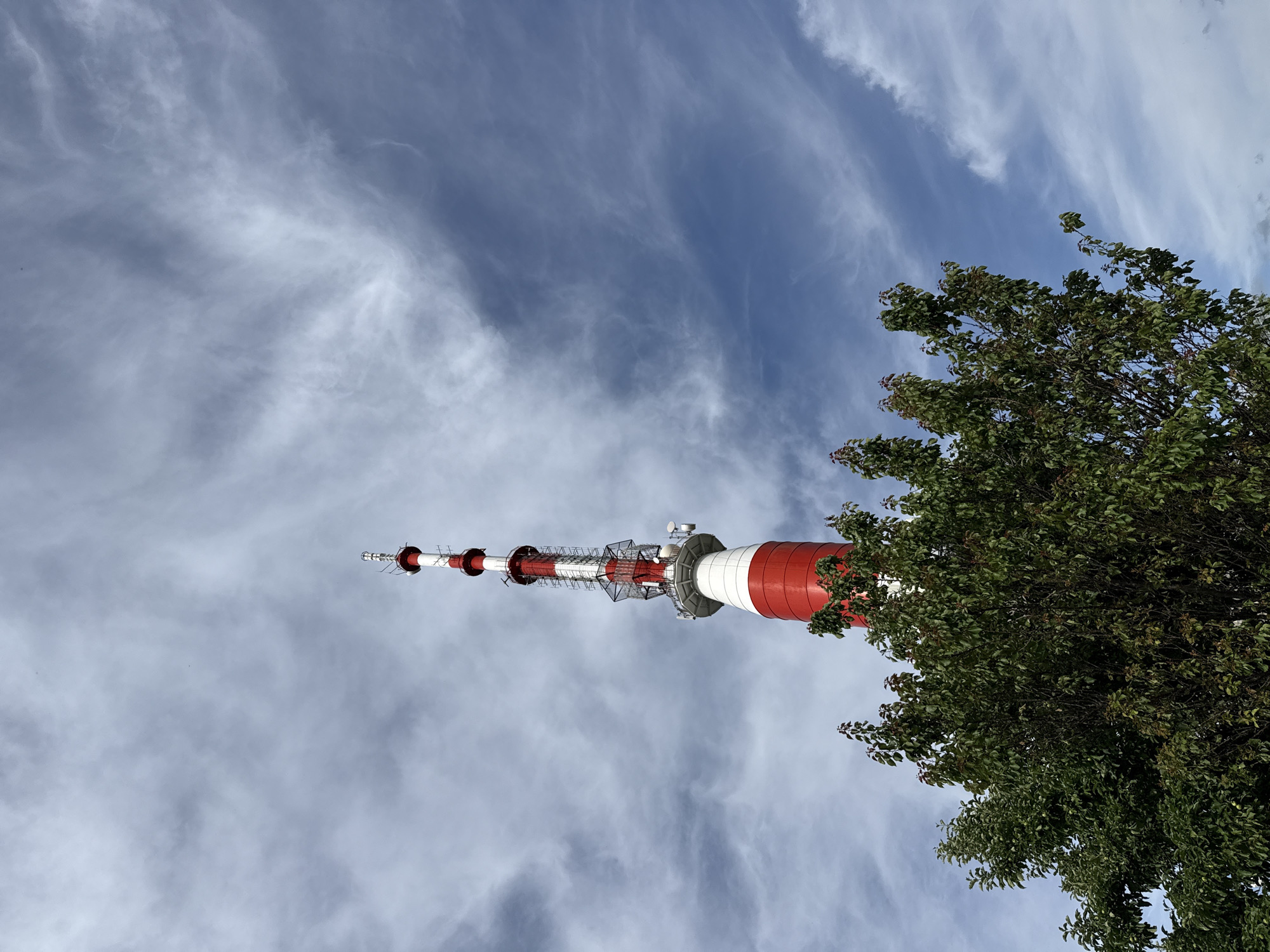 A red and white striped telecommunications tower stands tall against a cloudy blue sky. The tower is partially obscured by the lush green foliage of a tree in the foreground. The tower features several antennas and platforms at different levels. The clouds are wispy and spread across the sky, with some appearing more dense than others. The image appears to be taken from a low angle, looking upwards towards the tower. This structure is the Pleven Telecommunication Tower, located in the city of Pleven, Bulgaria.