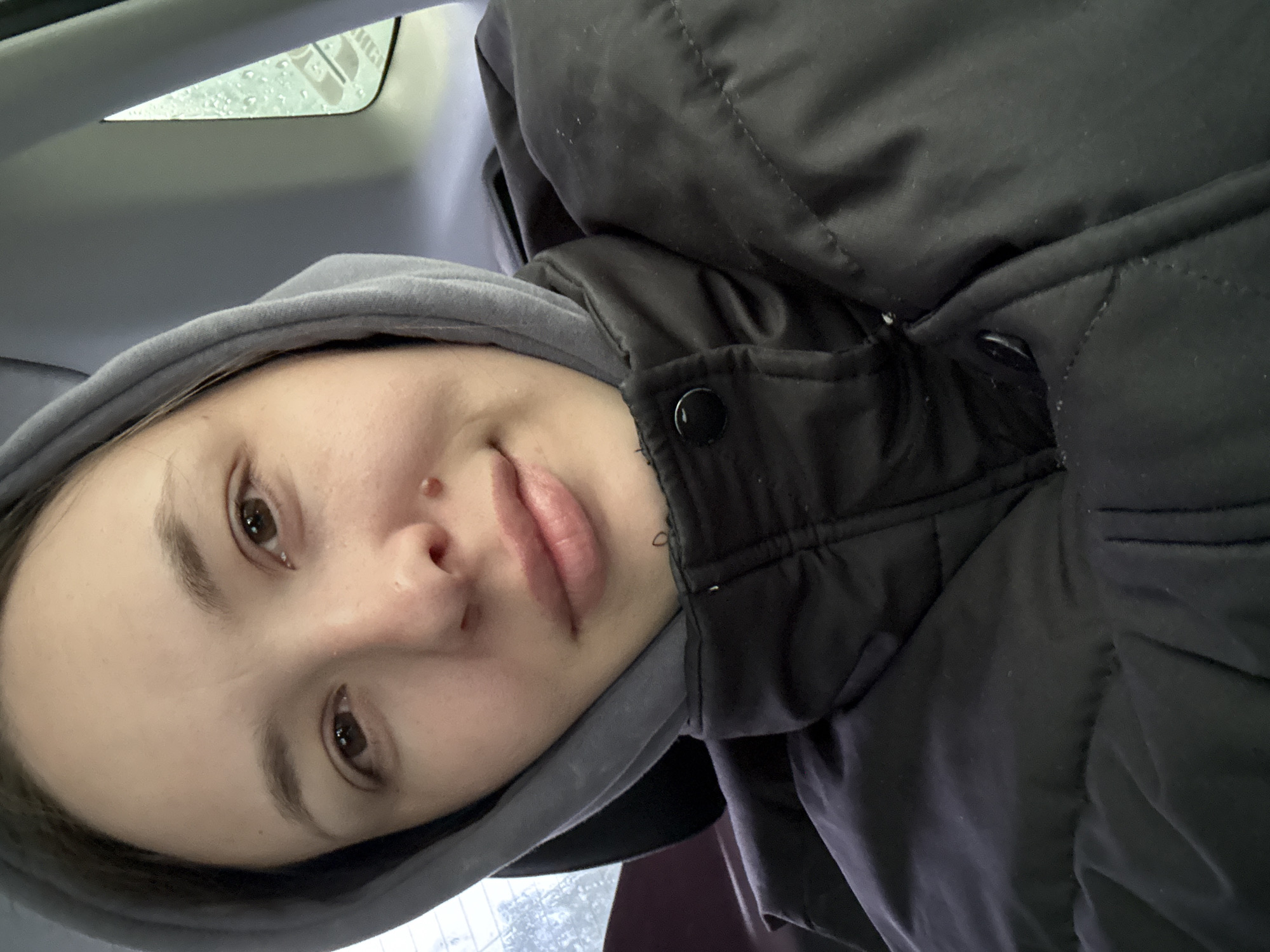 A young individual, likely female, is captured in a close-up, selfie-style portrait. She has fair skin, dark brown eyes, and a small piercing on her right nostril. Her expression is soft and relaxed, with a subtle, gentle smile. She wears a grey hooded sweatshirt underneath a black quilted puffer jacket, featuring visible snap buttons.
The setting appears to be inside a vehicle, possibly a car or bus, indicated by the grey plastic interior trim in the upper left corner and the window to her left. Outside the window, rain or condensation is visible, suggesting wet or damp weather in Chaikovsky, Russia. Through the blurred glass, a faint, possibly reversed white logo or text can be partially seen, appearing to contain the letters "HUG" or "HG" followed by "CH". The lighting is natural and soft, likely from an overcast or rainy day. The overall mood conveyed is calm and introspective.