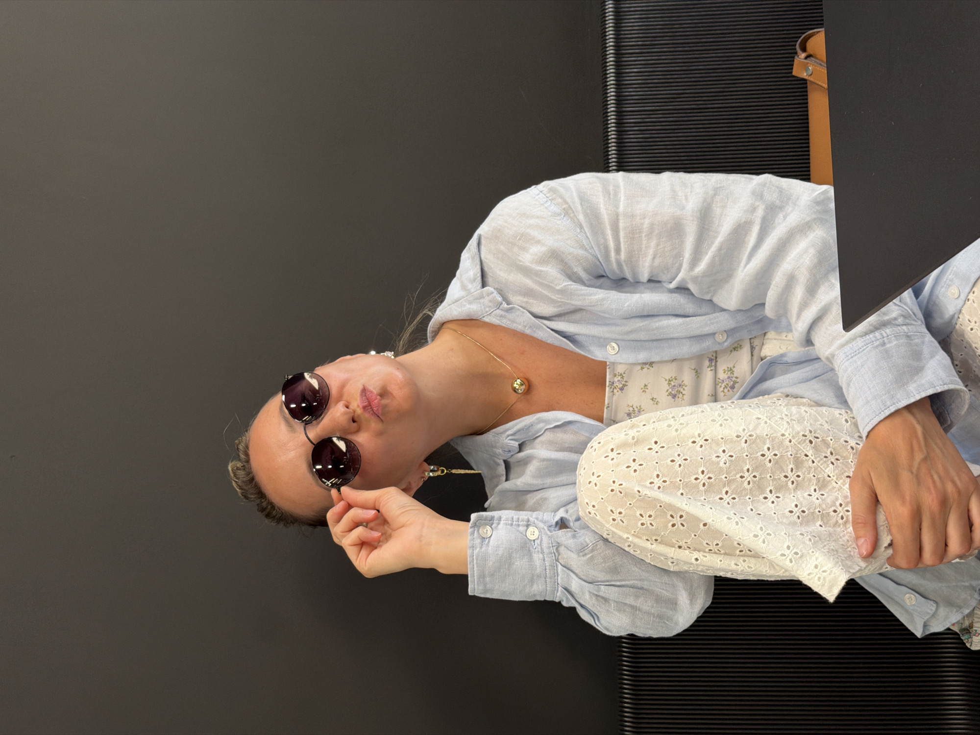 A woman with blonde hair pulled back in a bun wears round sunglasses, a light blue unbuttoned shirt over a floral top, and white eyelet pants. She is seated, holding her sunglasses with her right hand, and looking upwards with her lips pursed. A gold necklace with a spherical pendant is visible. The background consists of a dark wall and a black ribbed bench. A black laptop and a brown leather strap are on the right side of the frame. The image does not contain enough information to determine the location in the city of Izhevsk, Russia.