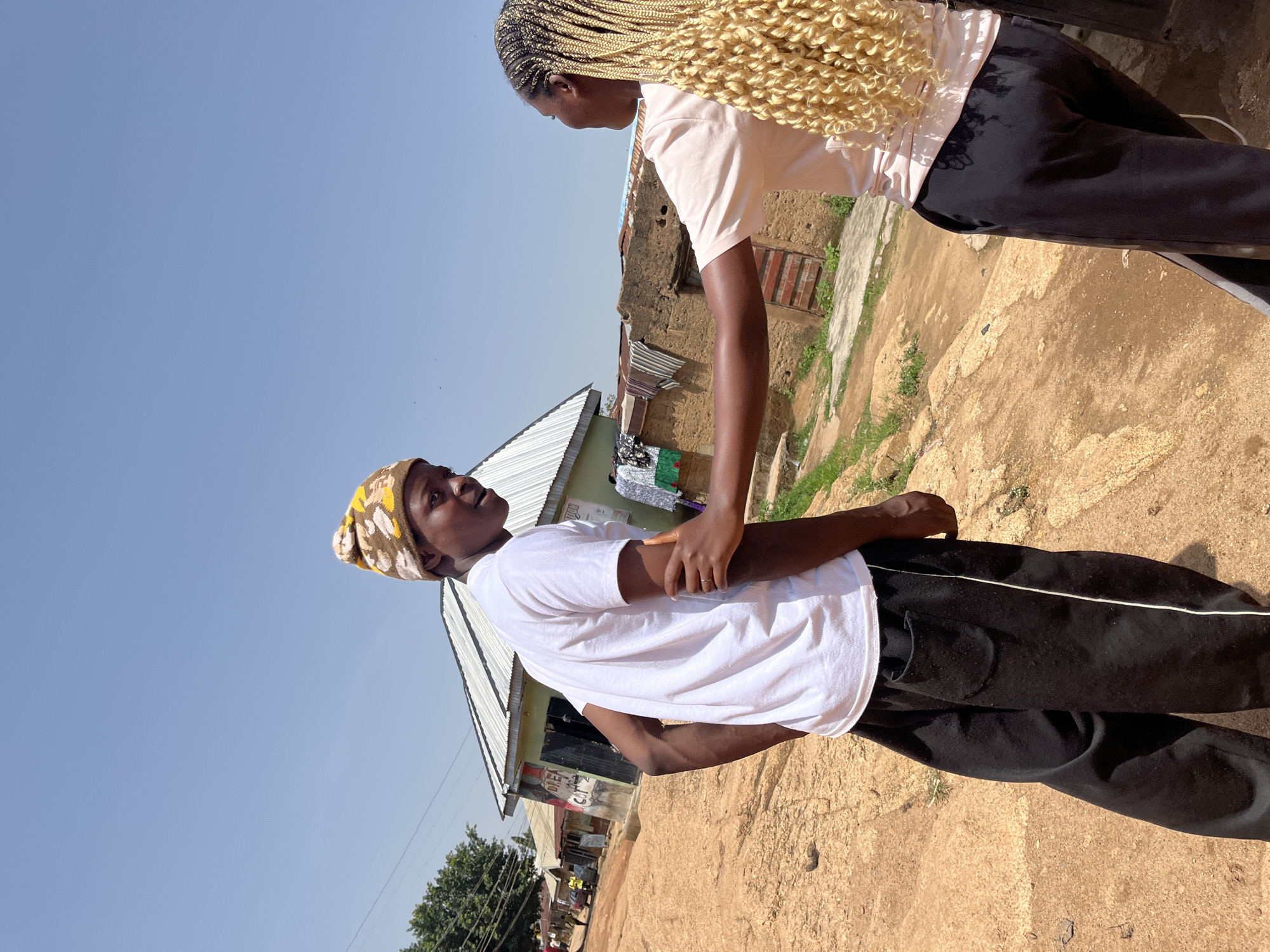 In Jos, Nigeria, two young individuals are engaged in an interaction on a dirt path. The person on the left, a young male, wears a white t-shirt, black track pants with a white stripe, and a patterned beanie. The person on the right, a young female, is seen from the back and right side, wearing a pale pink t-shirt and black pants. She has long blonde braided hair and her right arm is extended, touching the left person's arm. The background shows a typical urban setting with corrugated metal roofs on buildings, some with laundry hanging outside. The ground is uneven and dusty, characteristic of a dry season in Jos. The sky is clear blue, indicating a sunny day.