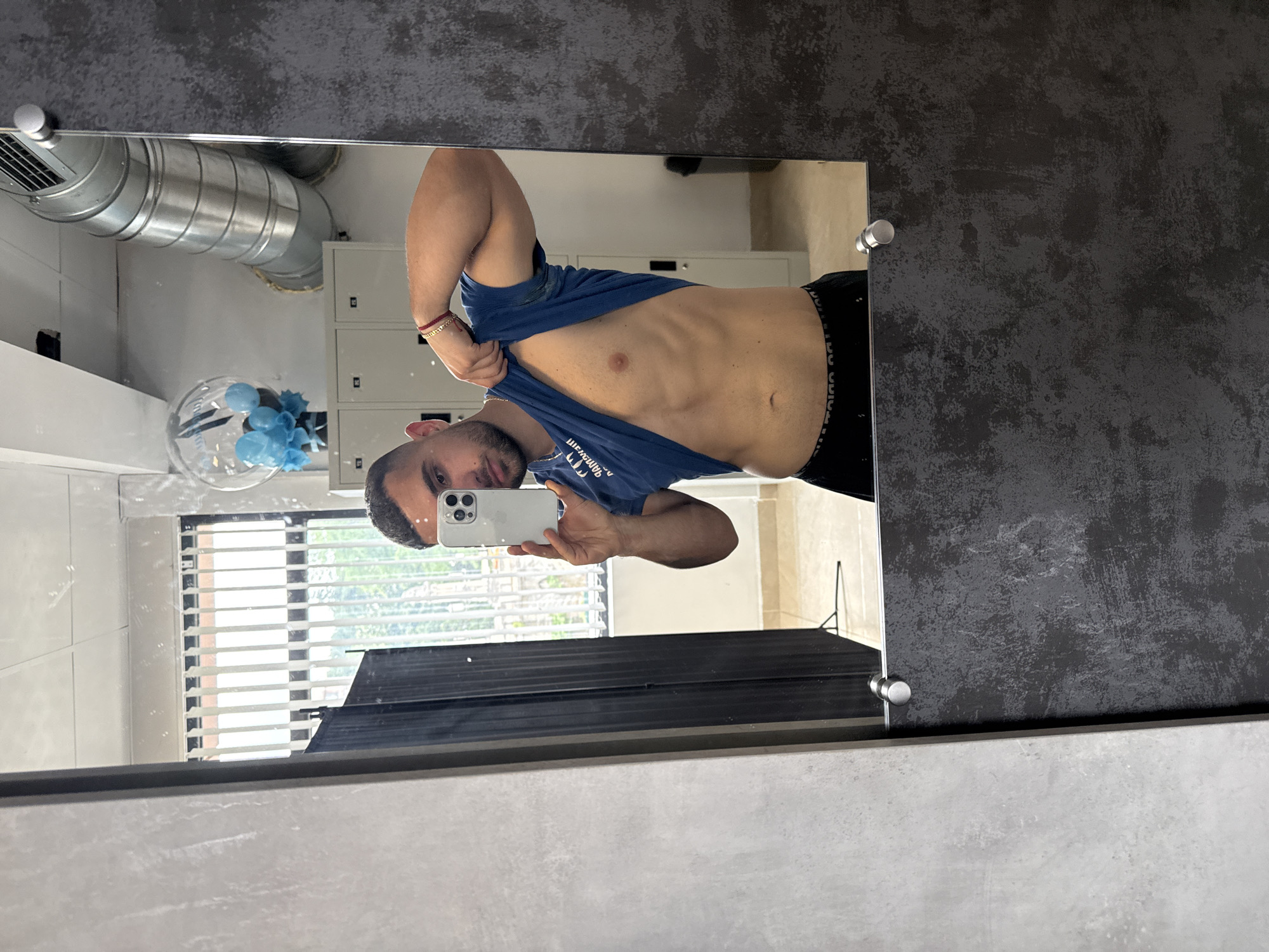A man is depicted taking a mirror selfie in what appears to be a gym or locker room. He stands facing the mirror, holding a silver-colored smartphone with a visible multi-lens camera array in his right hand. He has dark, short hair and a beard. The man is wearing a blue t-shirt, which he has pulled up with his left hand to expose his toned abdomen and navel. He also wears black underwear with a white elastic waistband. Notable accessories include a red and gold bracelet on his right wrist and a gold chain around his neck.
The background, reflected in the mirror, reveals several features of the setting. To the man's right, there are light-colored metal lockers. Above these lockers, industrial-style silver ventilation ducts run across the ceiling. In the lower left corner of the reflection, a clear glass sphere decoration containing light blue, ruffled flower-like elements is visible. Behind the man, a window with horizontal white blinds allows daylight to enter, with hints of greenery visible through the slats, indicating it is daytime. The walls of the room are a mix of light gray tiled surfaces and a darker, textured, possibly concrete or painted finish.
Visible text on the blue t-shirt, appearing mirrored in the reflection, reads "НИЗАГАМ ТРОПС," which when un-mirrored likely spells "СПОРТ МАГАЗИН" (Sport Magazine or Sport Shop in Cyrillic). The elastic waistband of his black underwear displays repeating white text or a logo, though it is indistinct. The mirror itself is framed by a dark, textured material on the top and sides, and a lighter gray material along the bottom, secured by cylindrical silver mounts.