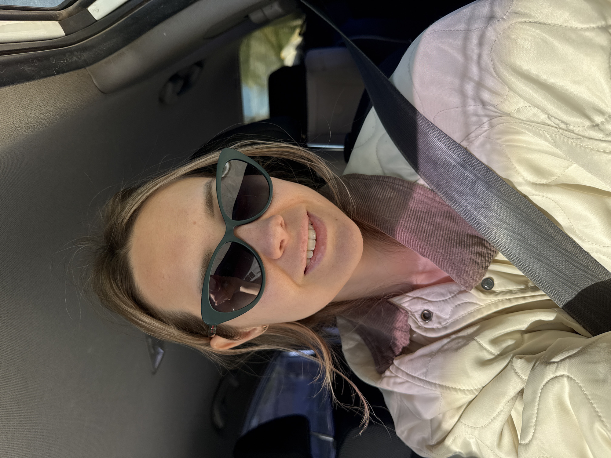 A woman wearing sunglasses and a white quilted jacket with a seatbelt across her torso sits inside a car. The interior of the car is visible, including the dashboard and ceiling. The location is unknown and cannot be confirmed as Chaikovsky, Russia from the information available in this image.