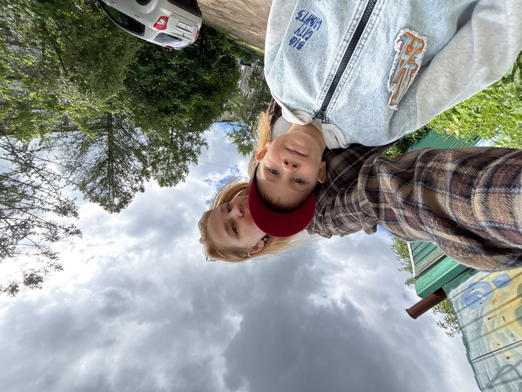 This outdoor selfie-style photograph, originally rotated, features two individuals – an adult and a child – against a backdrop of urban and natural elements under a cloudy sky.
In the foreground, a child with light hair looks directly at the camera with a neutral expression. They are wearing a light blue denim jacket over a grey hooded sweatshirt. The jacket features "BIG CITY LIGHTS" embroidered on one shoulder and an abstract orange and grey patch on the sleeve. Behind the child, an adult with blonde hair is partially visible, wearing a red baseball cap and a brown, beige, and white plaid shirt.
The background reveals a dirt road where a white car with Russian license plate "H748VC 67" is parked. Behind the road, numerous green trees and bushes lead to a multi-story apartment building, typical of post-Soviet architecture. To the right, a green corrugated metal fence or wall, adorned with blue and light-colored graffiti, has a brown pipe extending from it. The sky above is largely overcast with grey clouds, with some brighter patches visible.
The scene suggests a casual daytime moment, likely in a residential area of Rusanovo, Russia, as indicated by the location context and license plate. The weather is cloudy, and the overall mood is relaxed.