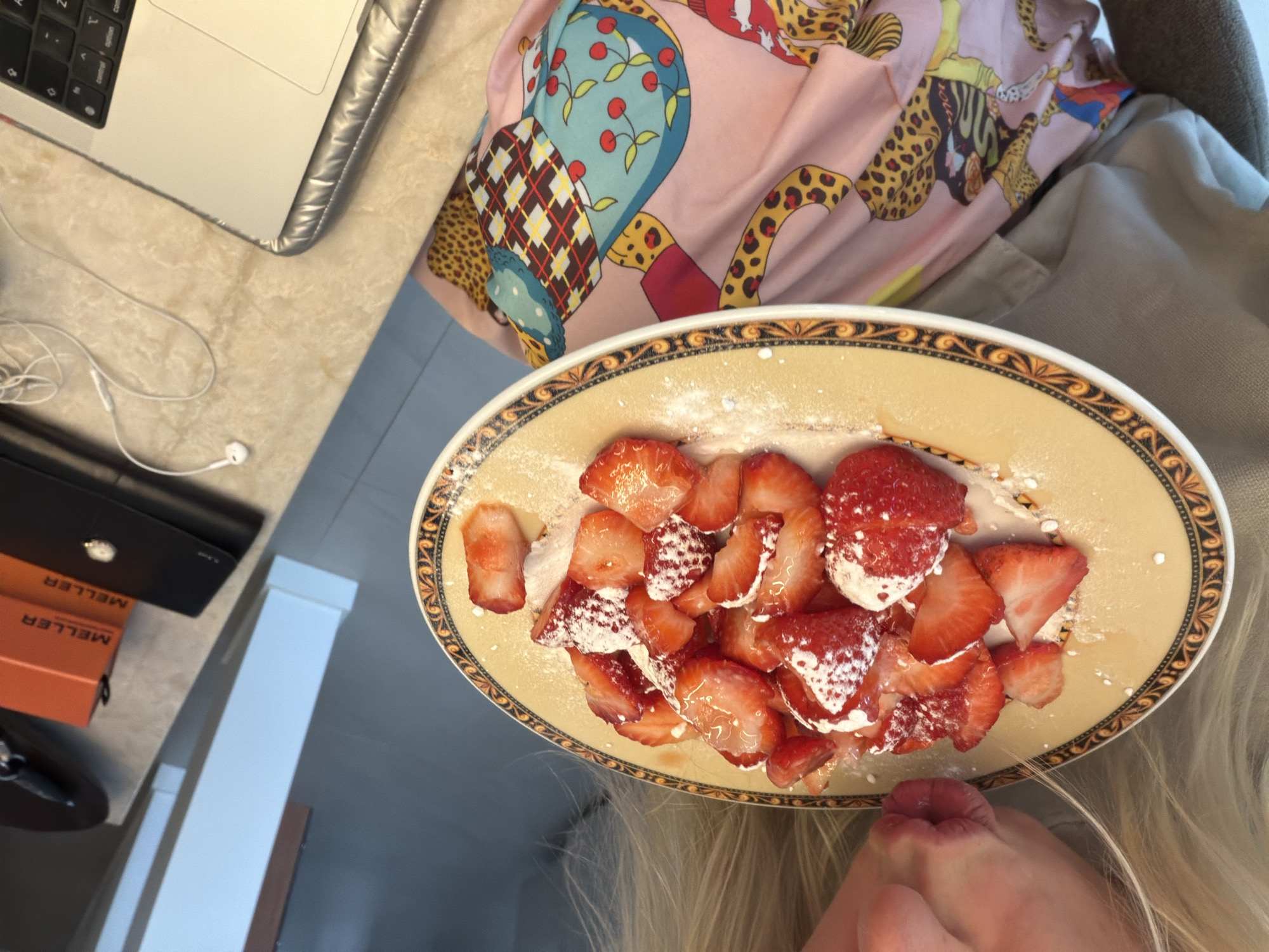 The image shows a close-up view of a person about to eat a plate of sliced strawberries dusted with powdered sugar. The plate has a decorative rim with an orange and brown pattern. The person's mouth is visible, pursed as if about to take a bite. They are wearing a light-colored garment and a pink fabric with a colorful animal print pattern. In the background, to the right, a laptop is partially visible on a countertop. Also on the countertop are two orange boxes labeled "MELLER" and a black wallet. A pair of white earbuds with a tangled cord lies next to the laptop. The environment appears to be indoors, likely a kitchen or dining area. The lighting suggests an indoor setting, possibly during the day. The exact location in Ridderkerk, Netherlands, is not discernible from the visual information provided.
