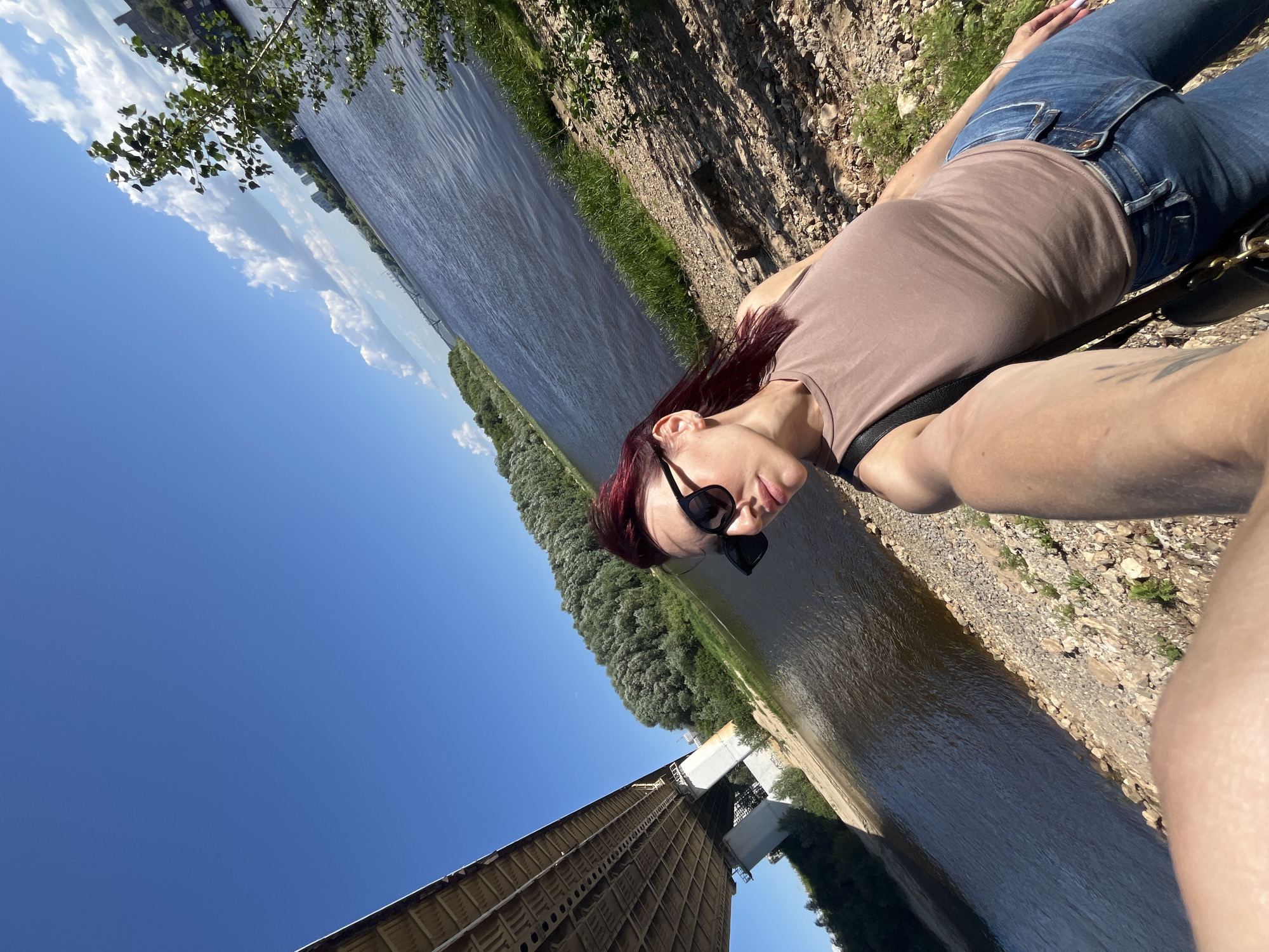 A woman with dark red hair and sunglasses takes a selfie on a sunny day next to a wide river. She is wearing a brown tank top and blue jeans. In the background, there is a large concrete bridge, a dense forest on the opposite bank of the river, and a clear blue sky with some white clouds. The location is the Nizhny Novgorod region, Russia, likely near the Volga River and one of its major bridges. The riverbank is rocky and dry, with some sparse vegetation.