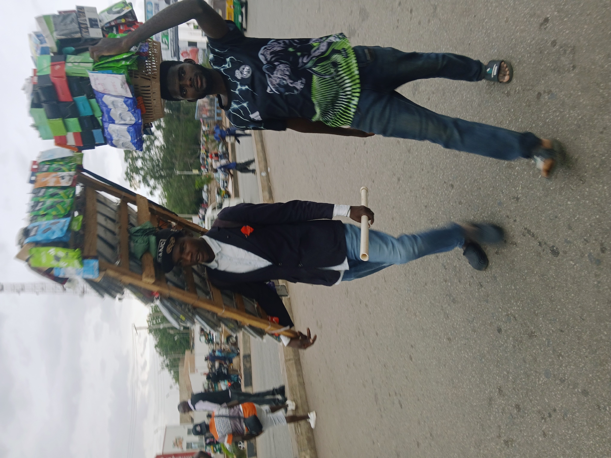 In Jos, Nigeria, two men are walking on a city street. One man carries a large stack of goods on his head with the help of a wooden frame. He wears a dark t-shirt with green accents and blue jeans, and is wearing sandals. The second man is wearing a dark blazer over a light shirt, with blue jeans, dark shoes, and carries a white pipe. The street has other people in the background.