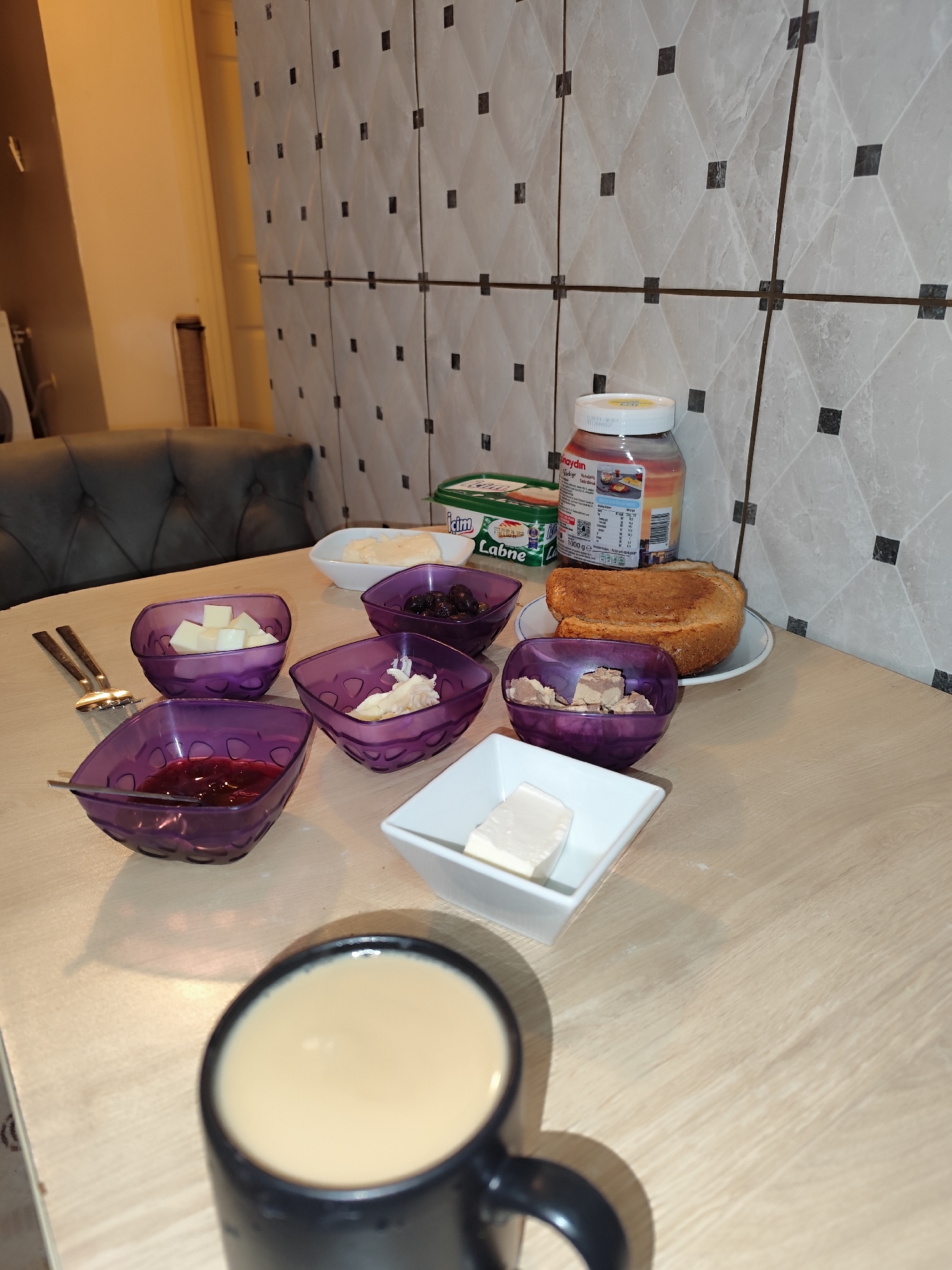 The image depicts a breakfast table in a likely residential setting in Sarıyer, Turkey. The table has a light wood grain surface. On the table are several purple square bowls containing: cubed cheese, olives, a white spread, what appears to be chopped liver or pâté, and jam. There's a white rectangular dish with butter, a white round dish with slices of white cheese, a green package of "Labne" cheese, a jar of "Tunaydin" hazelnut spread, and a plate with two slices of toast. A black mug filled with a beige liquid, probably coffee or tea, is in the foreground. Silverware is near the cheese cubes. The background features a tiled wall pattern. In the left background, a portion of a grey sofa can be seen. The overall scene suggests a typical Turkish breakfast spread.