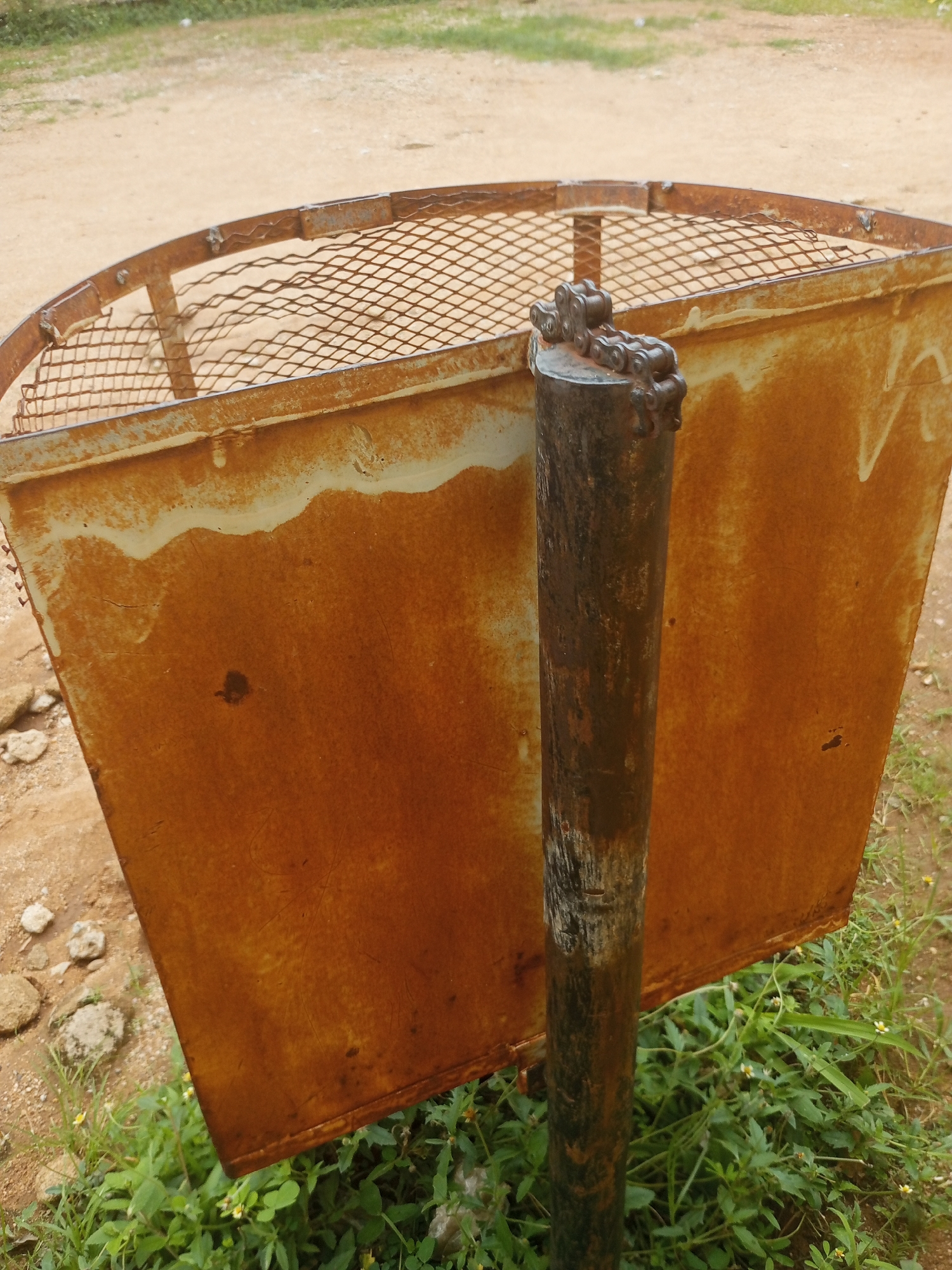 A rusty metal trash receptacle stands outdoors on a patch of dry earth and green vegetation. The trash can has a wire mesh top rim and a solid, Rusted metal body with some lighter colored streaks. A dark, rusty metal pole supports the trash can from the front, with a segment of a metal chain secured to the top of the pole. The background consists of dry, dusty ground and more green foliage. The image is likely taken in Jalingo, Nigeria.