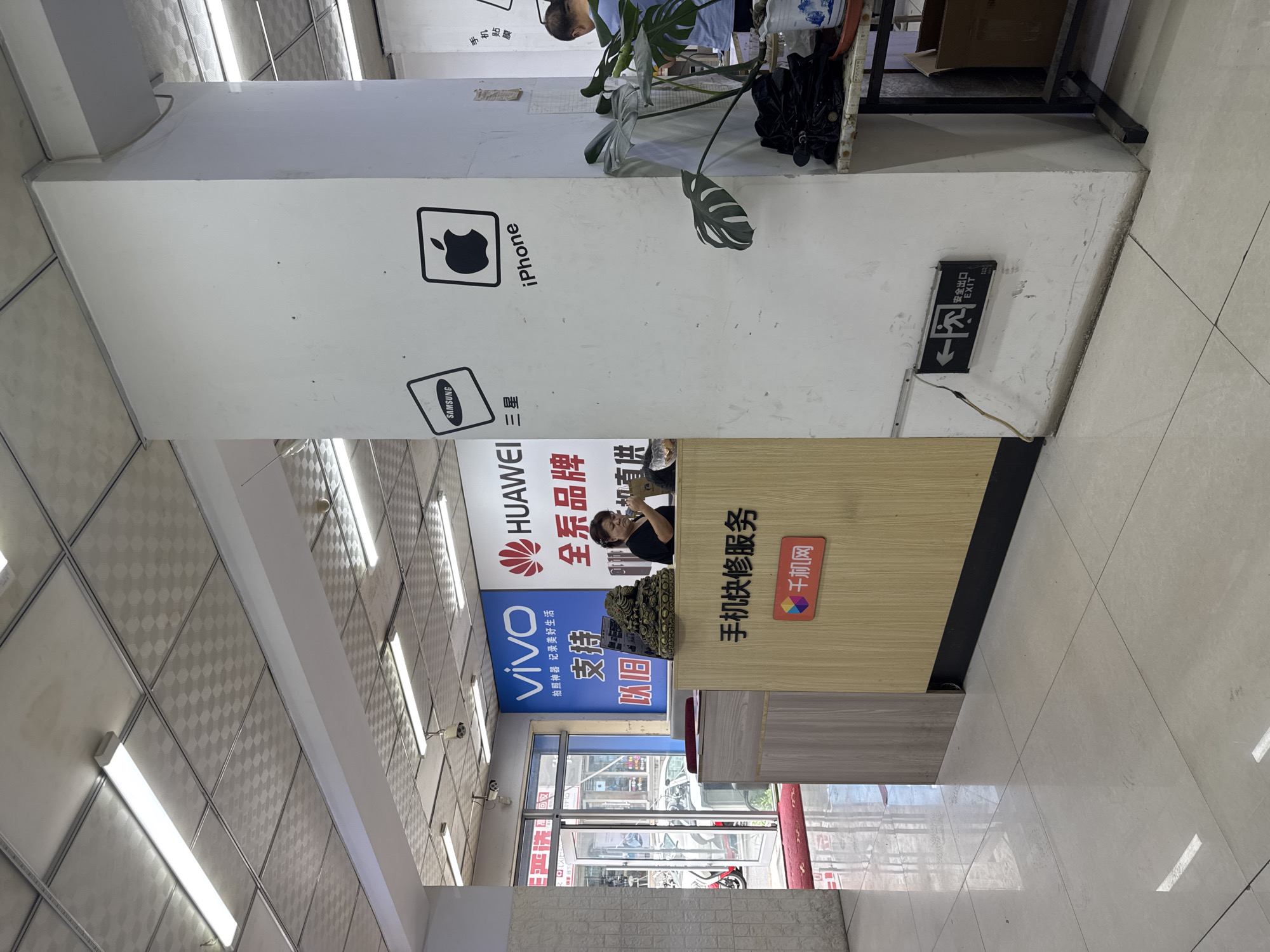 This image depicts the interior of a mobile phone repair shop located in Dadong District, China. The scene is dominated by a white pillar that prominently displays logos for Samsung and iPhone. A counter is situated in the foreground, behind which a woman is seen interacting with a customer. The counter itself has signage in Chinese characters, including "手机快修服务" (Mobile Phone Repair Service) and a logo with "千机网" (Qianji Network). In the background, a large blue and white sign advertises "VIVO" and "HUAWEI" phones. The overall environment appears to be a retail space with fluorescent lighting on a tiled ceiling. An exit sign with an arrow pointing left is visible on the lower part of the white pillar.