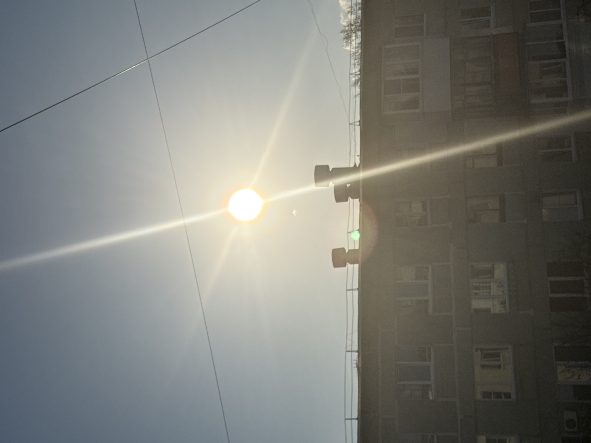 The image shows a view of a tall building in Chaikovsky, Russia, likely an apartment block, under a bright, sunny sky. The sun is positioned prominently in the center, creating a lens flare effect with prominent rays emanating from the center. Electrical wires cross the sky. The building is situated on the right side of the frame, displaying multiple windows that suggest it is a multi-story structure. Some sort of ventilation system is present on the building. The sky is clear and pale blue, creating a high-contrast image due to the intense sunlight.