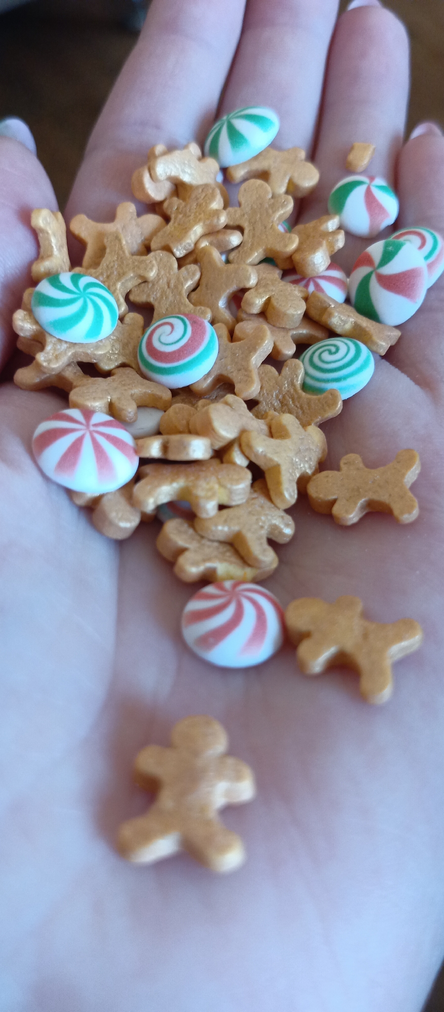 A hand is holding a collection of candy sprinkles. The sprinkles include small, golden-colored gingerbread men and round, white candies with green and red spiral patterns. The background is blurry. The image does not provide information about location.