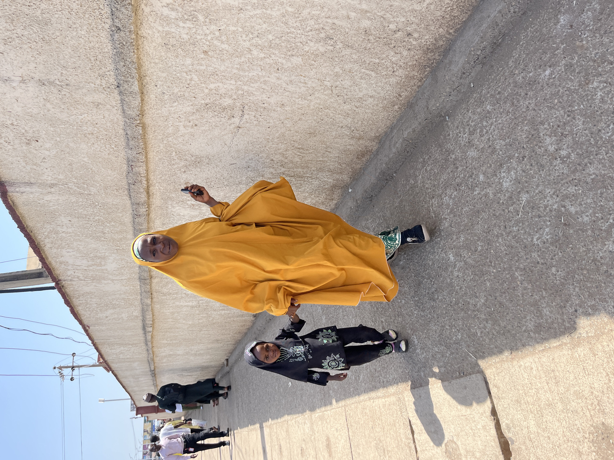 A woman in a yellow hijab and a child in a black outfit with light-colored embellishments are walking down a paved street in Jos, Nigeria. The woman is holding a black object in her right hand. Several other people walk in the background on the same side of the street. The street is bordered by a high, gray concrete wall on one side and a low building and power lines on the other, with a clear blue sky visible above.
