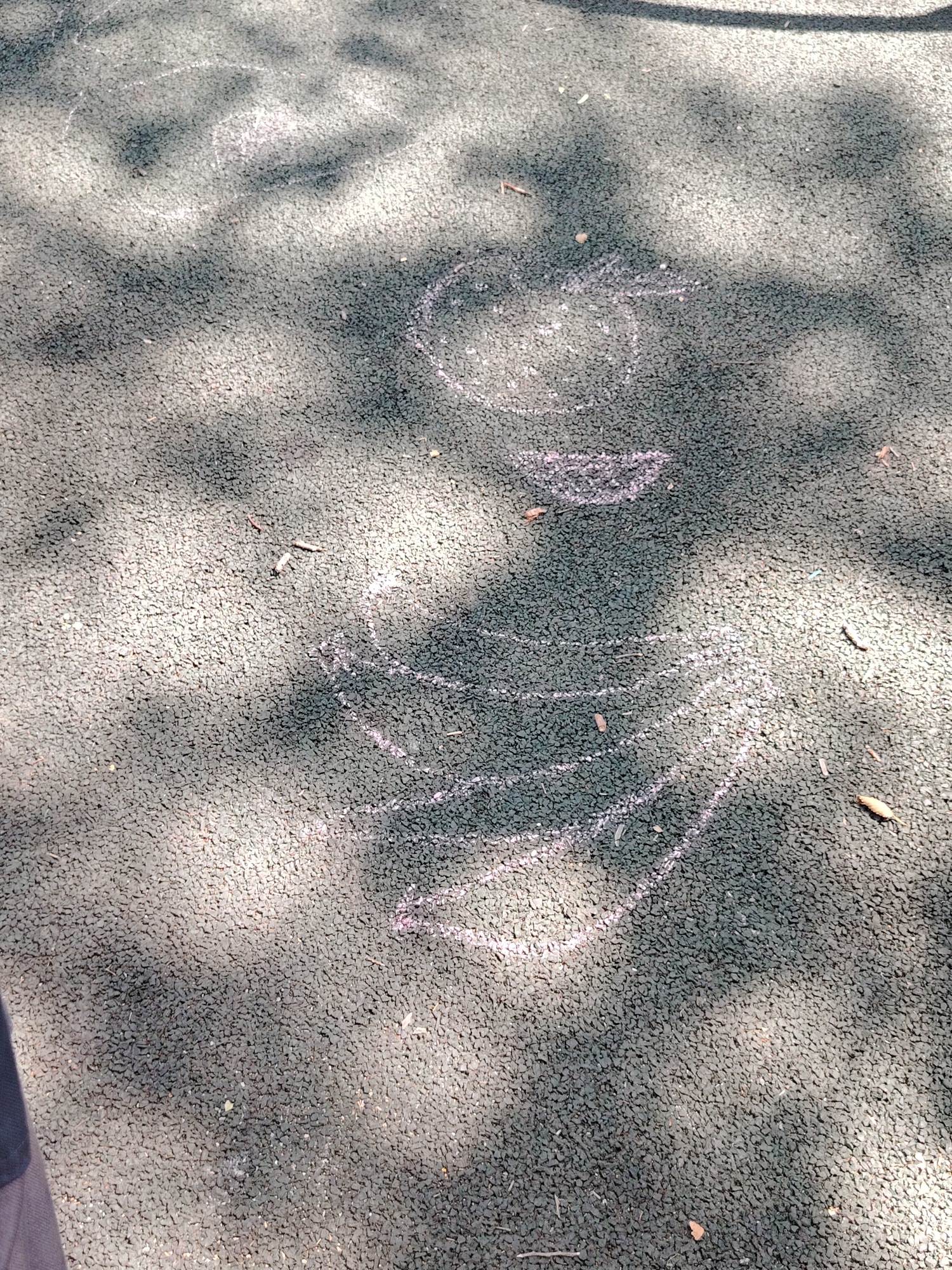 The image shows a chalk drawing on a dark, textured outdoor surface, possibly a playground. The drawing appears to be a stylized figure or object with curved lines, some of which are pink and some white. The surface is mottled and shows dappled sunlight and shadow. There are a few small twigs and dry leaves scattered on the surface. The image does not contain any information that would allow for the identification of its location within the city of Astana, Kazakhstan.