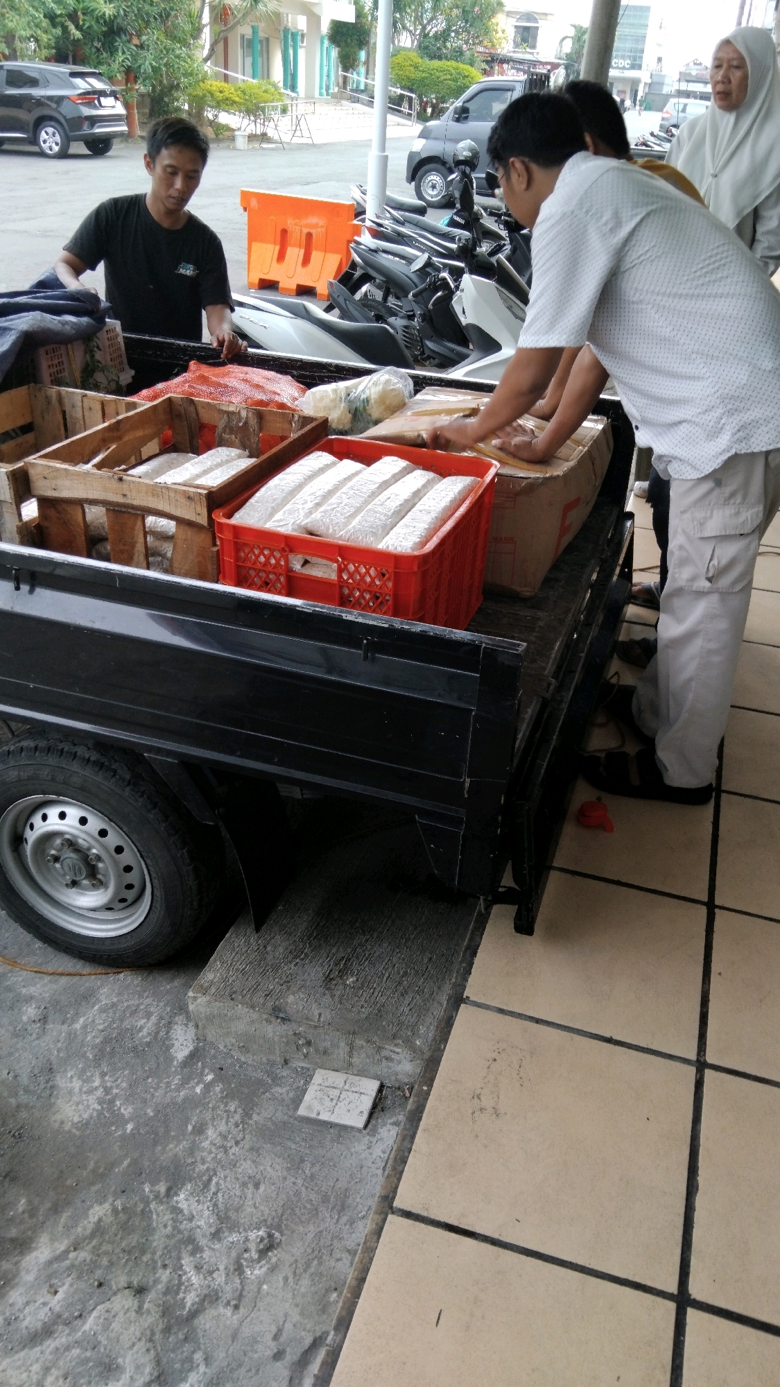 In a street scene in Pekalongan, Indonesia, a black pickup truck is parked next to a tiled walkway and a concrete surface. The truck bed contains several items, including wooden crates filled with what appear to be packaged food items, and an orange plastic crate holding more of these packaged items. A man in a black t-shirt is leaning into the truck bed, reaching towards the contents. To his right, another man wearing a white patterned shirt and light-colored pants is interacting with the items in the truck bed. Behind them, a woman wearing a white hijab is standing, observing the scene. In the background, several motorcycles are parked, and various vehicles are visible on the street. The overall environment suggests a commercial or delivery setting in a moderately urban area.