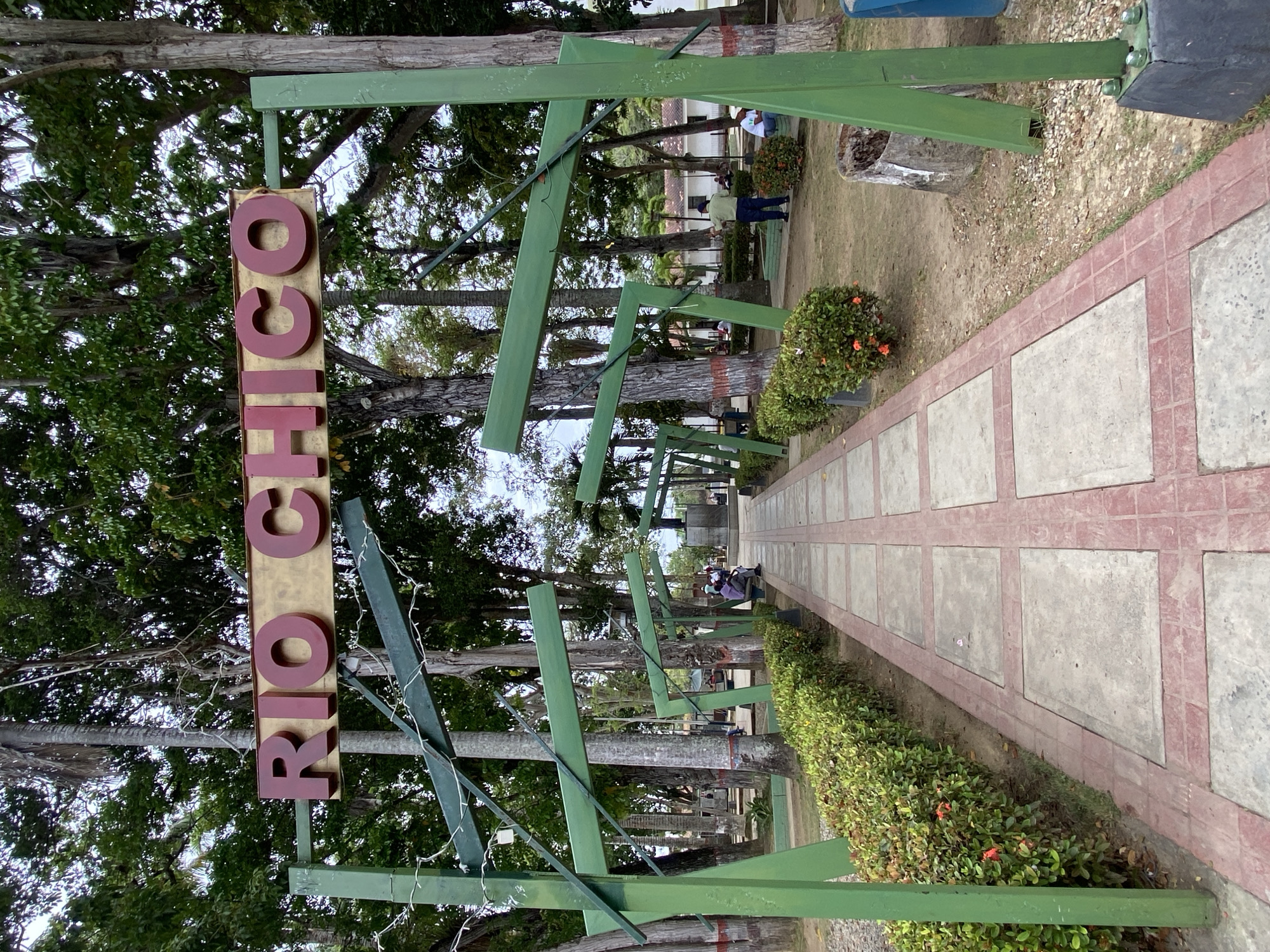 The image shows a pathway in Río Chico, Venezuela. The path is lined with greenery and decorated with green wooden structures. The sign "RÍO CHICO" stands at the entrance. Several people are visible in the background. The environment is lush with trees. The sky is overcast.