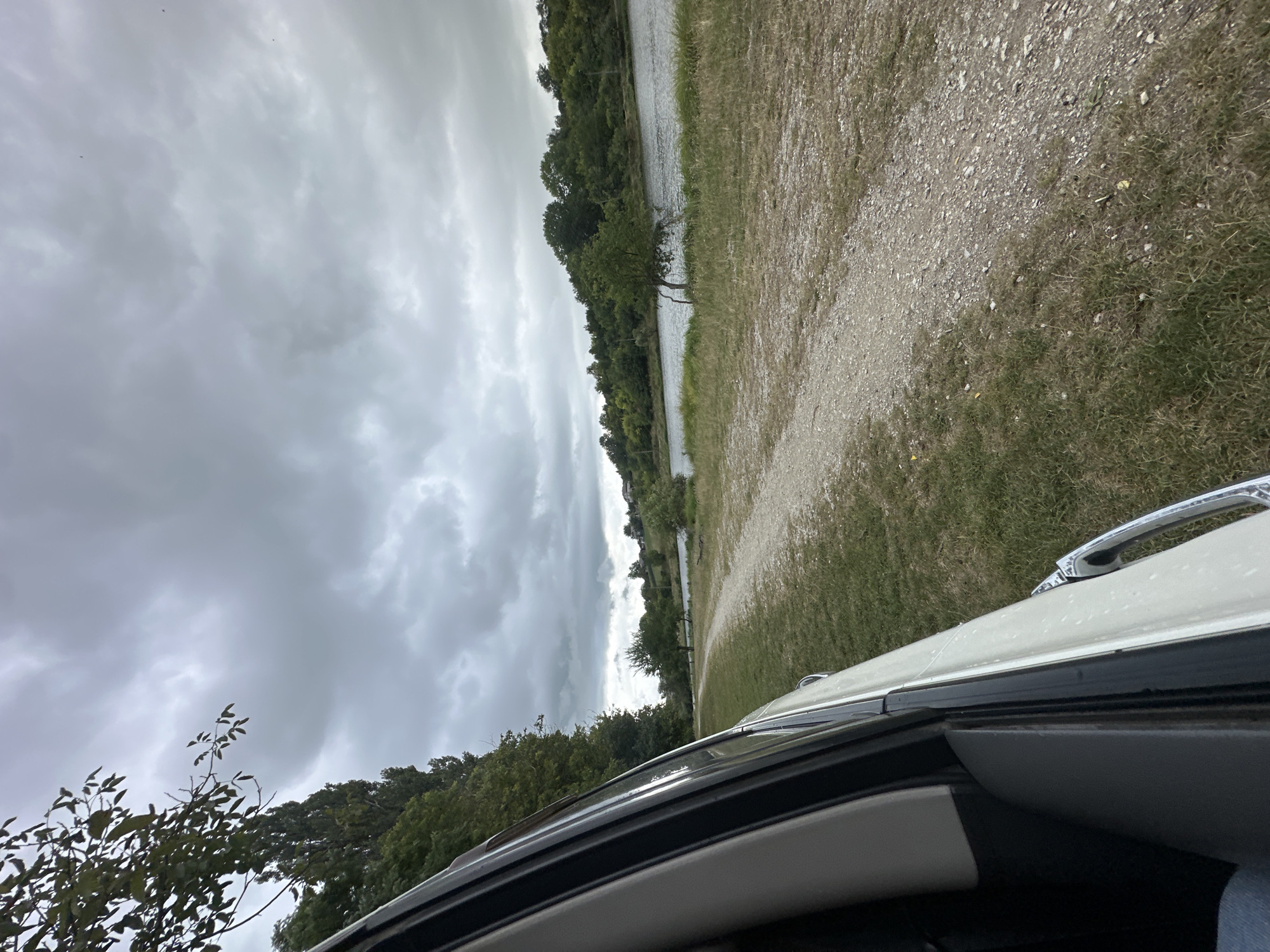 An outdoor, eye-level, medium shot captures a view from the passenger side of a light-colored vehicle looking out towards a tree-lined riverbank under a cloudy sky. The vehicle's side and window frame are visible in the foreground, taking up the left portion of the frame. A dirt path extends from the foreground into the midground, curving to the right along the edge of a grassy area. The river flows in the midground, bordered by a densely wooded hill on the far bank. The sky is heavily overcast with dark, dramatic clouds. The scene depicts a natural, possibly rural setting, with no people or animals clearly visible. The location is identified as Makiv, Ukraine.
