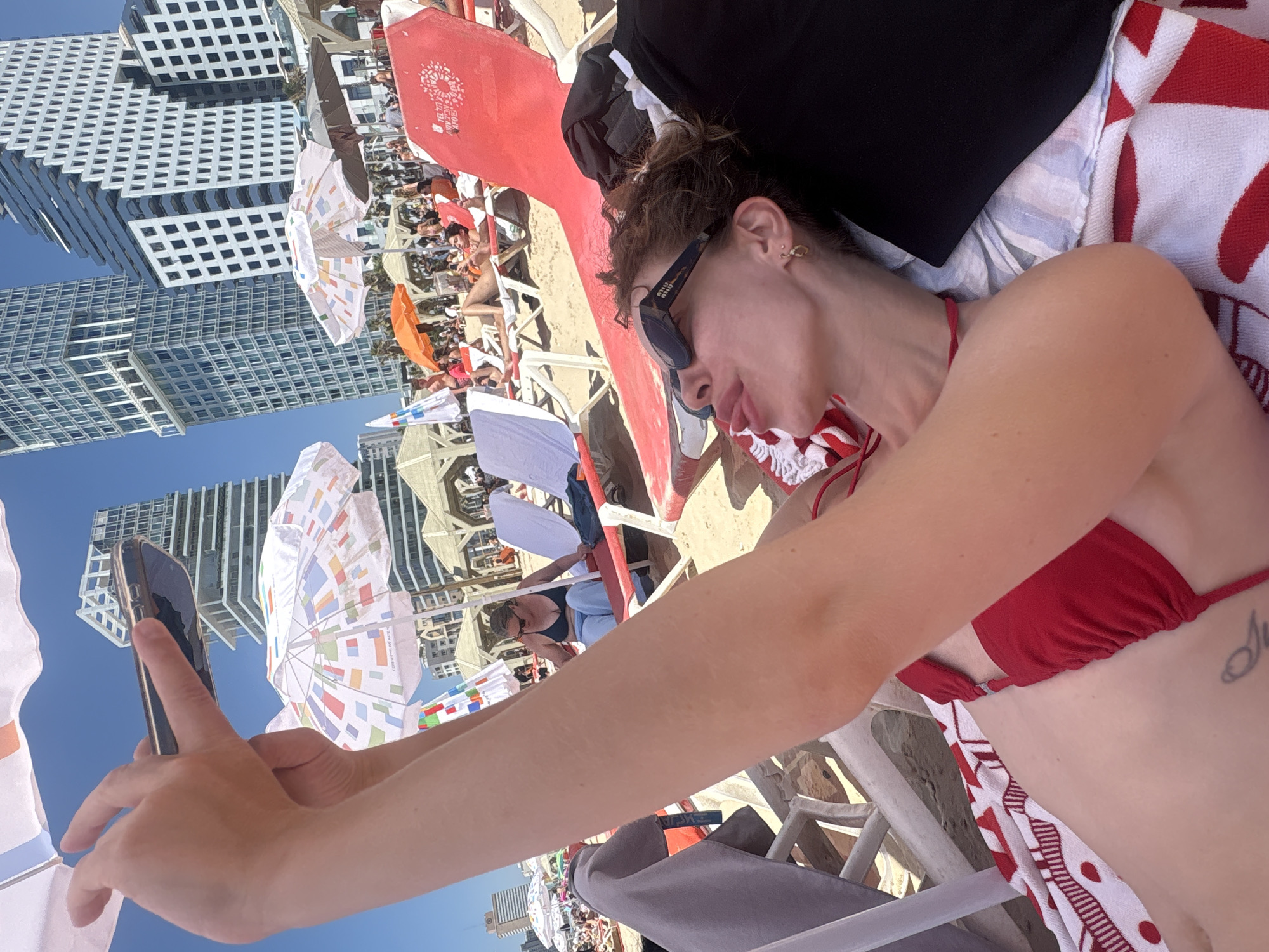 The image depicts a woman lying on a beach chair on a sunny day, taking a selfie with her phone. She is wearing a red bikini and sunglasses. In the background, there are other beachgoers, umbrellas, and modern skyscrapers, indicative of the Tel Aviv skyline. The beach appears to be crowded, with many people relaxing and enjoying the weather. The focus is on the woman and her activity, with the urban environment serving as a backdrop.