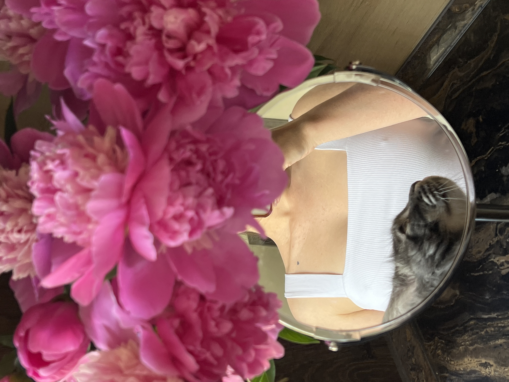 A high-angle shot shows a mirror on a dark, patterned countertop, reflecting a woman in a white tank top and a grey cat. The woman's upper chest and shoulder are visible, and she is holding a bouquet of large, pink peonies, which obscure the top of her head and face. The cat, seen from the side, appears to be sniffing something in the foreground. The background is blurred, but suggests a domestic setting. The image was taken in Ternopil, Ukraine.