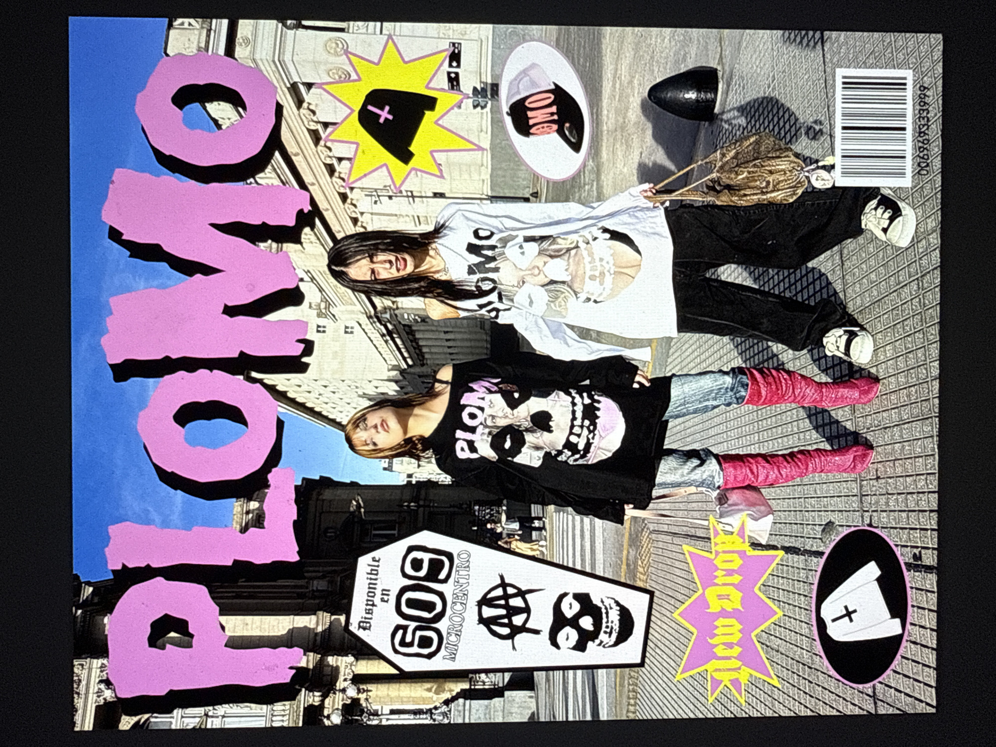 The image shows a magazine cover titled "PLOMO" with two female models posing on a city street. The location is Buenos Aires, Argentina. One model wears a black long-sleeved shirt with a skull graphic and pink, knee-high boots. The other model wears a white long-sleeved shirt with a graphic and black pants. The background includes urban architecture, with buildings and sidewalks visible. Additional design elements on the cover include graphic symbols such as a coffin with the number "609" and a skull, stars, a barcode, and the words "New Project."