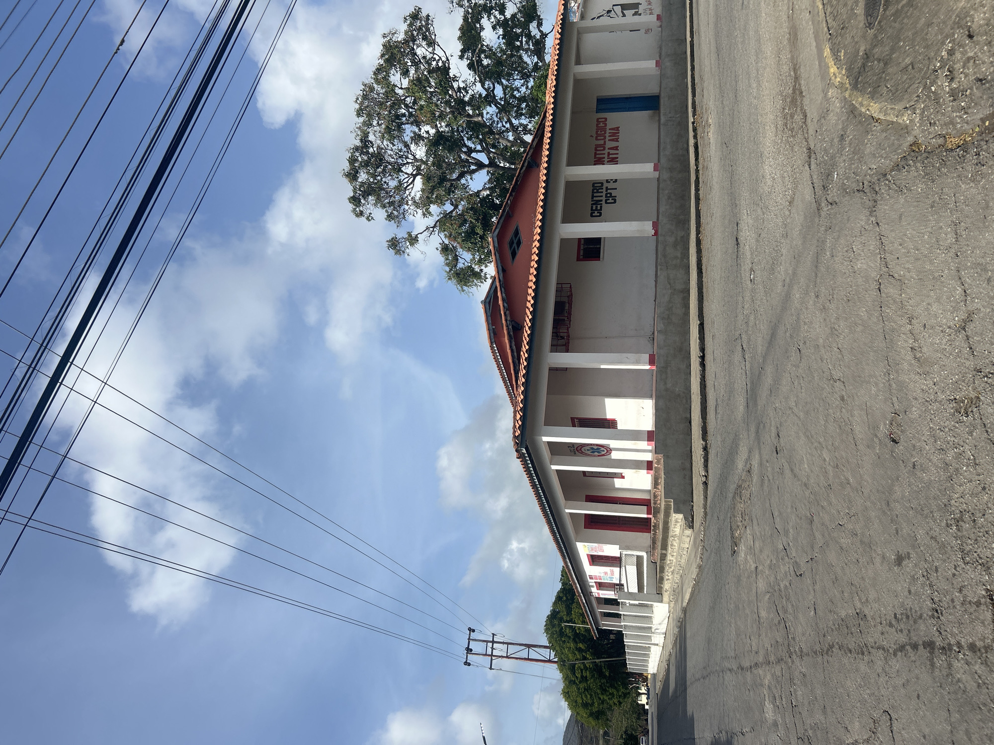 The image shows a one-story building with a red tiled roof located on a corner in Santa Ana, Venezuela. The building is painted white with red trim around the windows and pillars. Signage on the building reads "CENTRO ODONTOLOGICO CPT 3 SANTA ANA". A blue door is visible on the right side of the building. The building is situated on a paved road that appears to be in disrepair, with cracks and potholes visible. Overhead power lines crisscross the sky, supported by a utility pole. Trees and a hillside are visible in the background. The sky is partly cloudy.