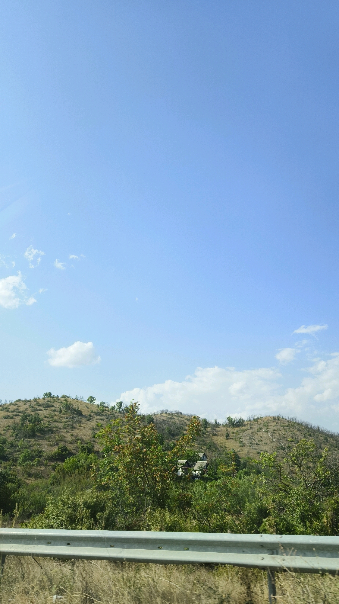 A wide, vertical shot captures a landscape under a clear, bright blue sky with sparse white clouds. In the foreground, a grey metal guardrail runs horizontally across the bottom of the frame. Beyond the guardrail, dry, light-brown grasses are visible.
The middle ground features a rolling hillside densely covered in vegetation. The hillside exhibits varied shades of green, with some areas appearing dry and brown, interspersed with patches of vibrant green trees and bushes. Scattered throughout the greenery, two houses with white walls and dark roofs are nestled into the hillside.
The background consists of the expansive blue sky, taking up approximately two-thirds of the image. The scene is a rural landscape, likely in the vicinity of Badar, North Macedonia, given the hilly terrain and vegetation. No people or animals are visible.