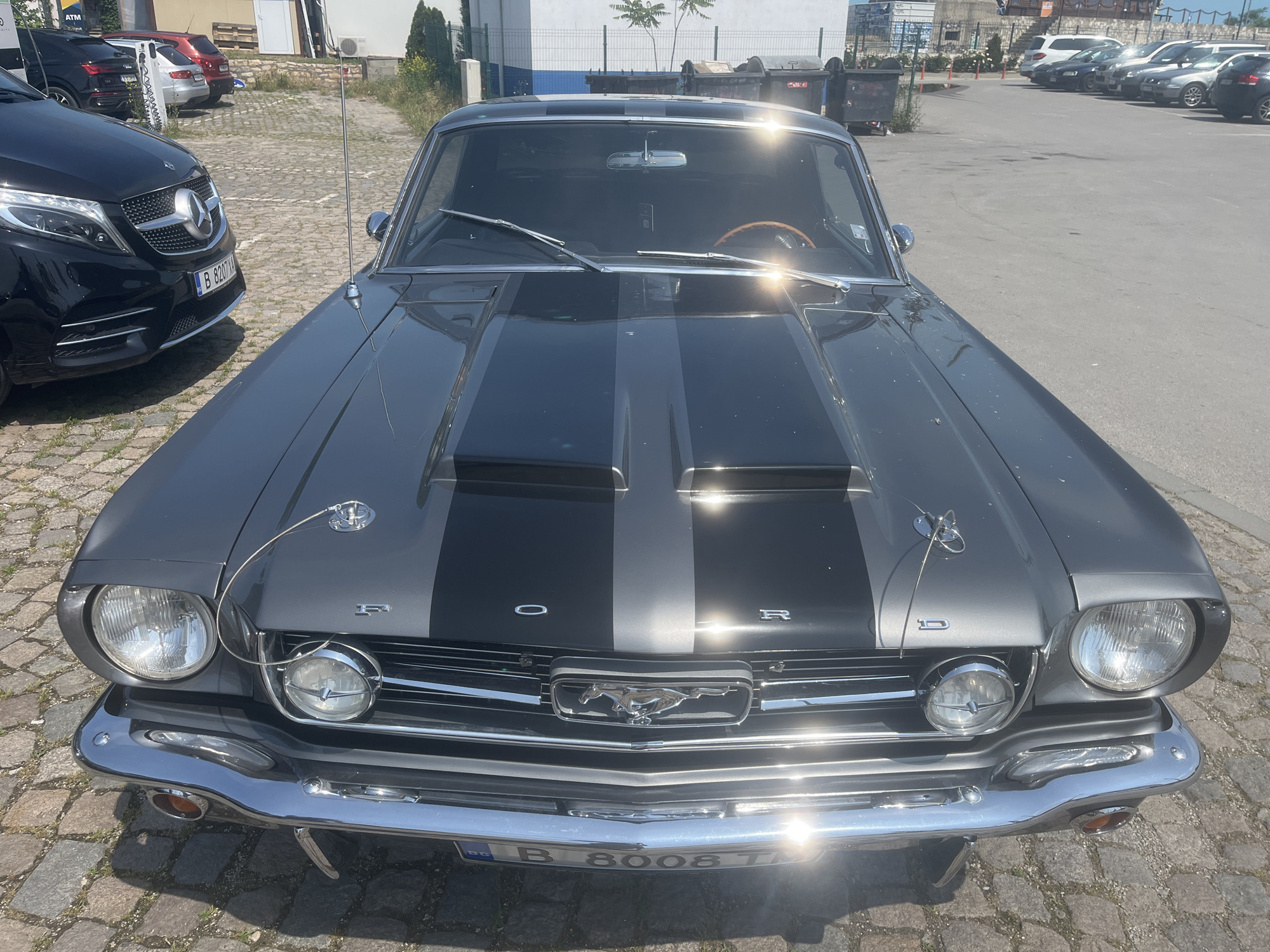 This media file presents a front view of a classic dark grey Ford Mustang, parked outdoors on a sunny day. The car features two wide black racing stripes running down its hood, which also bears the letters "FORD" spelled out in white. The Mustang has prominent chrome details, including its front bumper, grille, headlight bezels, and fender emblems. Circular auxiliary lights are positioned below the main headlights, and hood pins secure the front of the hood. An antenna extends from the front fender. Through the windshield, a wooden steering wheel is visible. The license plate on the Mustang reads "B 8008 T," indicating Bucharest, Romania.
To the left, partially visible, is a modern black Mercedes-Benz van with its own license plate, "B 8207 M." In the background, numerous other cars are parked in what appears to be a mixed-surface parking lot, with cobblestones in the foreground and asphalt further back. Buildings, an "ATM" sign, and several large dumpsters are also visible in the distance. The bright sunlight creates strong reflections on the glossy surfaces of the cars, particularly on the Mustang's hood and windshield. The scene is static, depicting vehicles parked under a clear, sunny sky.