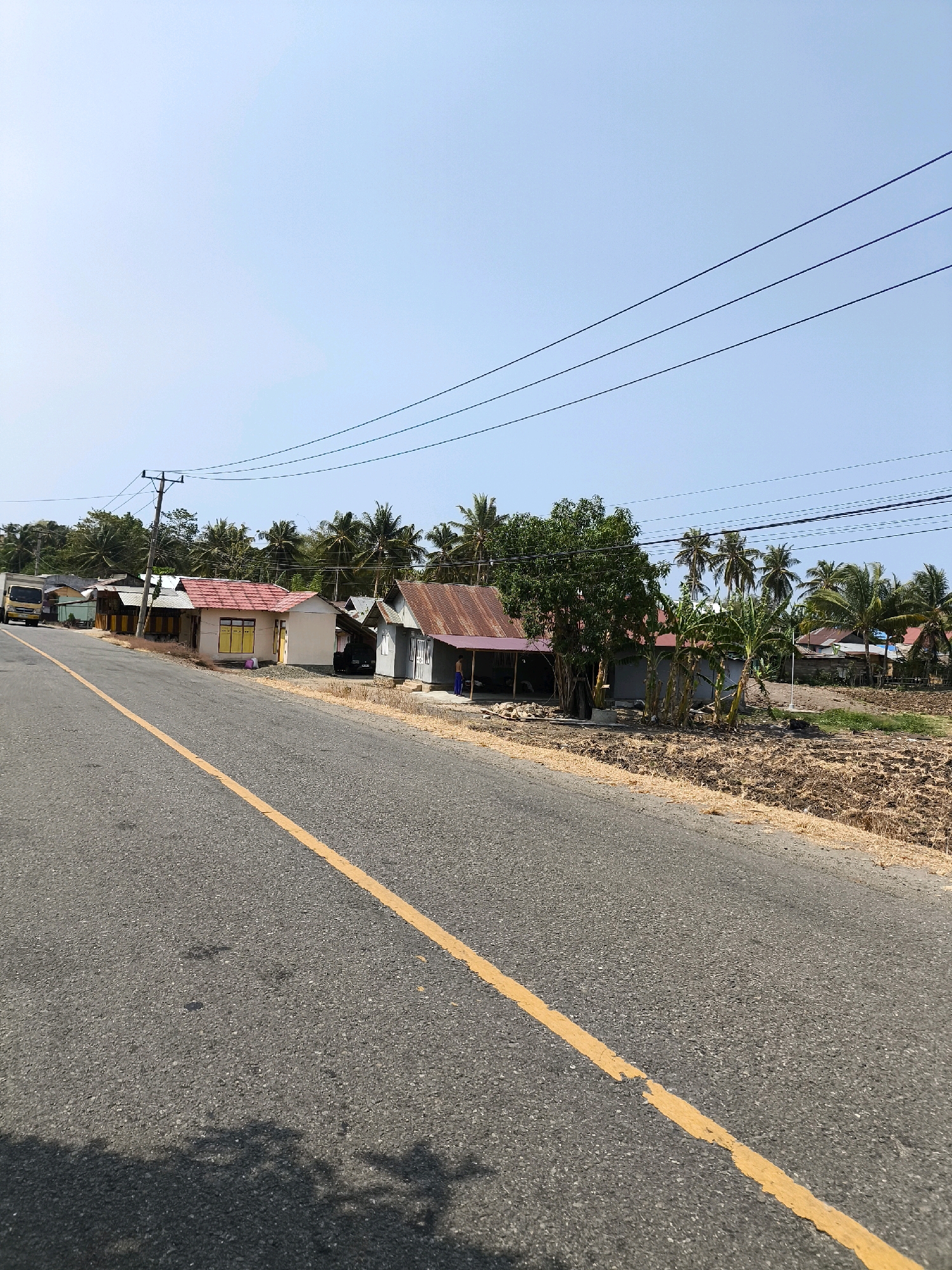 A rural road in Kwandang, Indonesia, stretches into the distance under a clear blue sky. The asphalt road features a broken yellow dividing line. On the right side of the road, several simple, modest houses with corrugated tin roofs are visible, some with painted walls and yellow window frames. Beyond the houses, a line of palm trees and other vegetation forms a backdrop. The land in front of the houses appears to be dry and cultivated, with visible tilled soil and scattered dried grasses. A yellow truck is parked on the left side of the road, further down the street, near a row of structures. Overhead, several power lines are strung between utility poles.